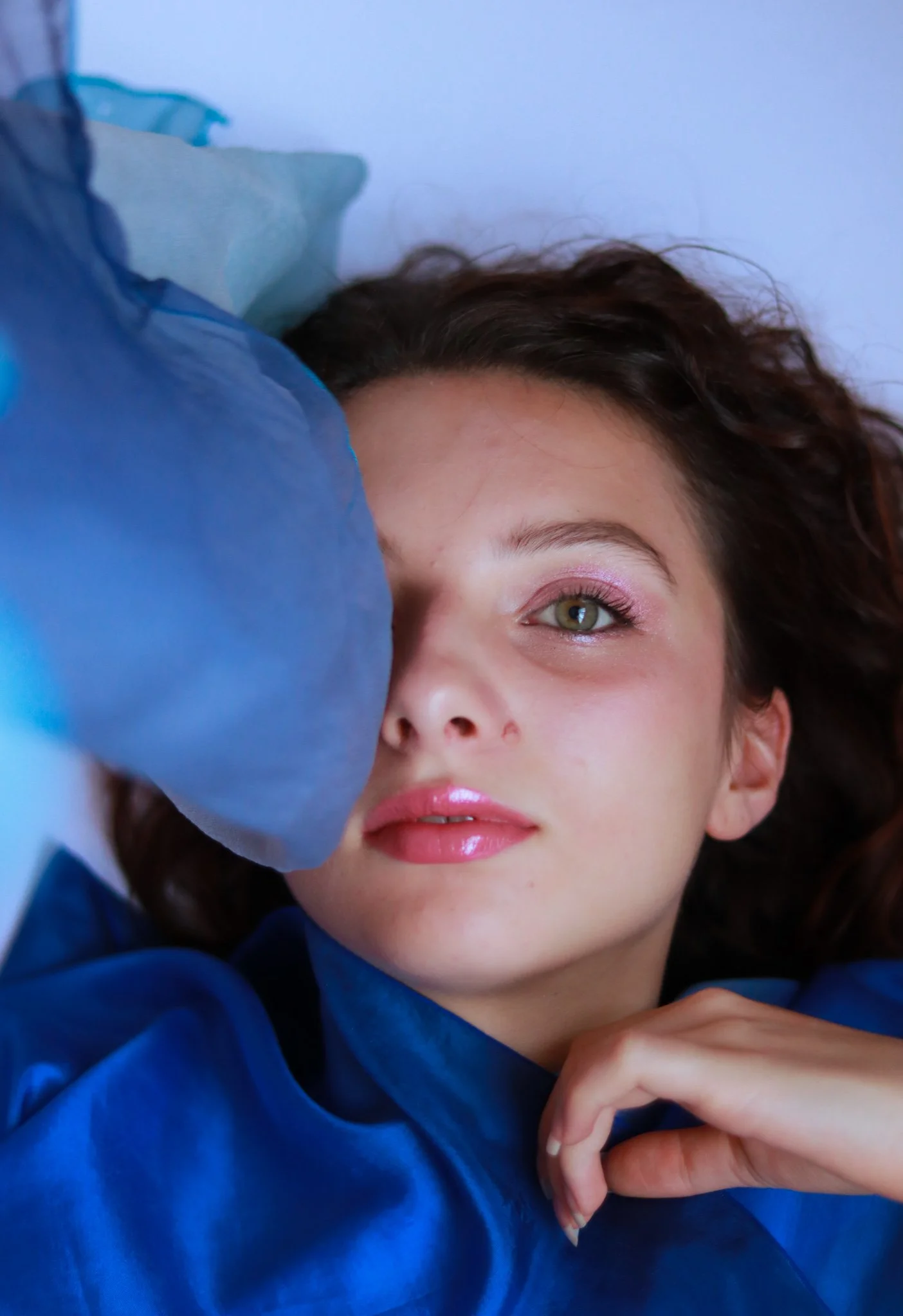 Une jeune femme aux cheveux bouclés bruns, portant un vêtement bleu, repose sur le sol ou un lit, regardant la caméra d'un œil charmant et détendu.