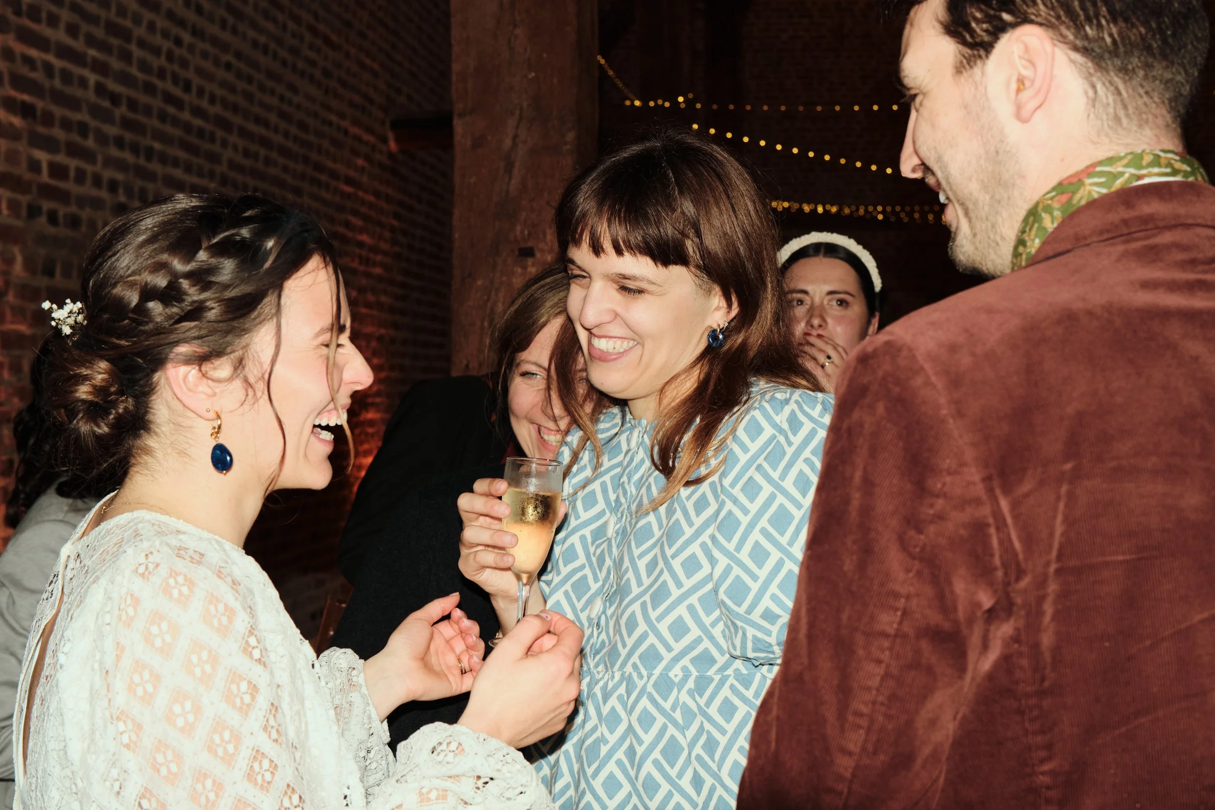 Un groupe de personnes souriantes lors d'une célébration, échangeant des regards heureux, avec une femme tenant une flute de champagne. Contexte festif avec guirlandes lumineuses.