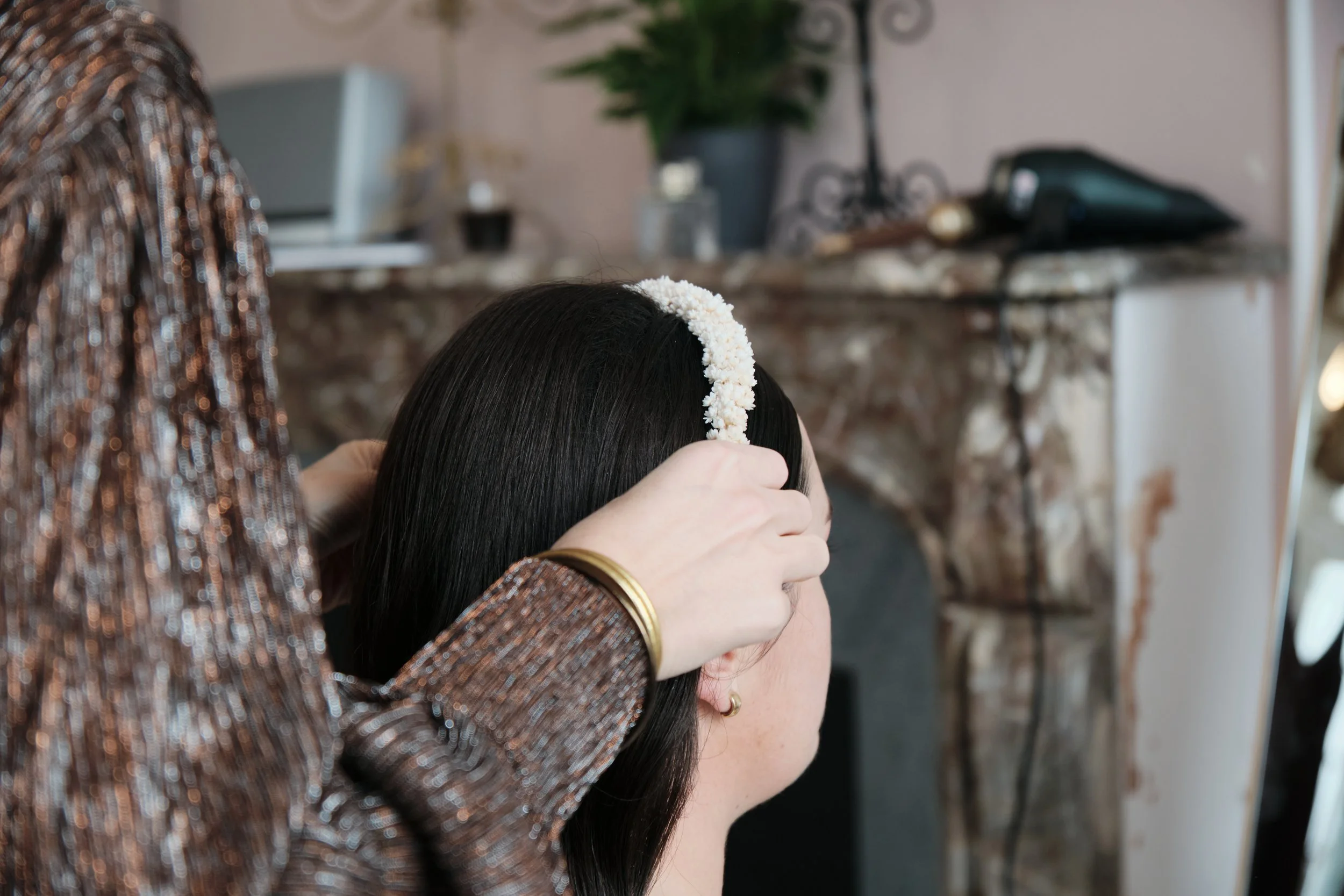 Une femme coiffe une autre femme, qui porte un bandeau de fleurs sur la tête, dans un intérieur avec cheminée en marbre et décorations diverses.