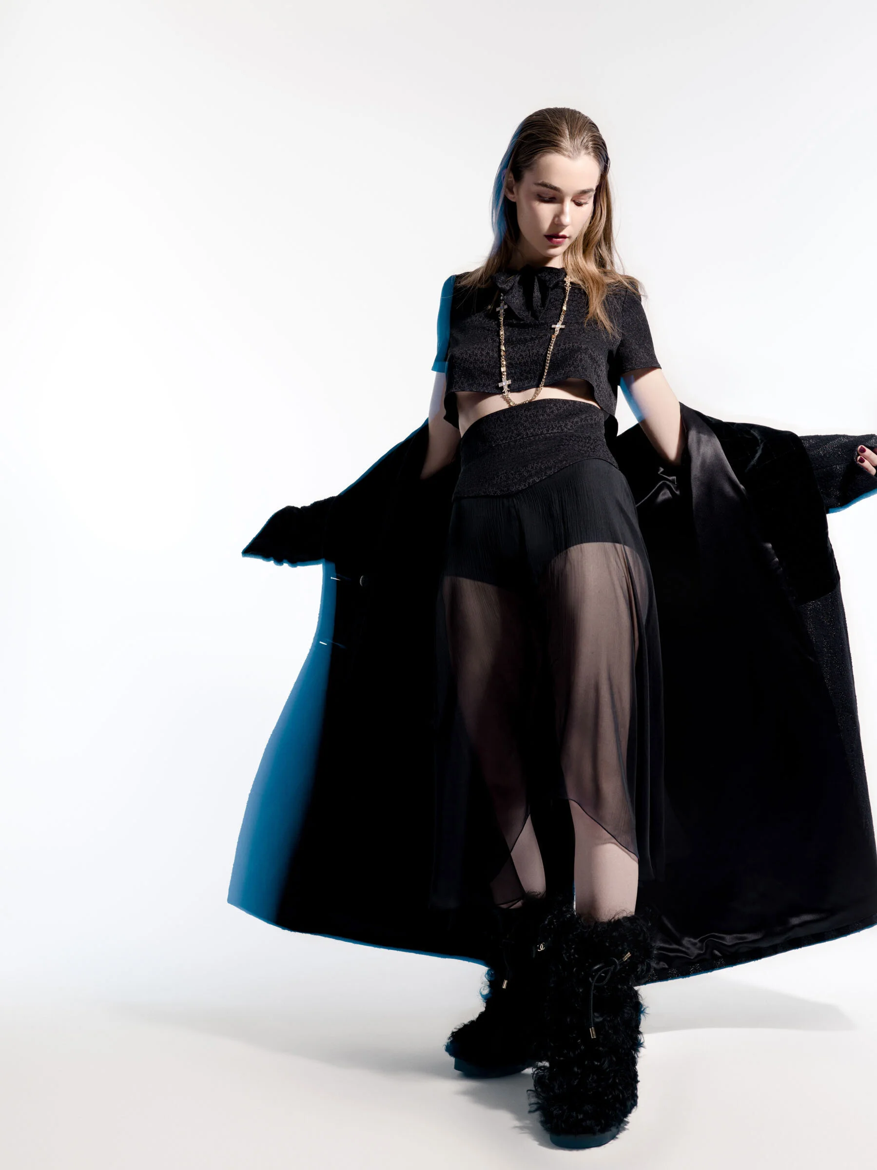 Young woman wearing black sheer skirt, cropped top, long coat, and fuzzy boots, posing against a white background.