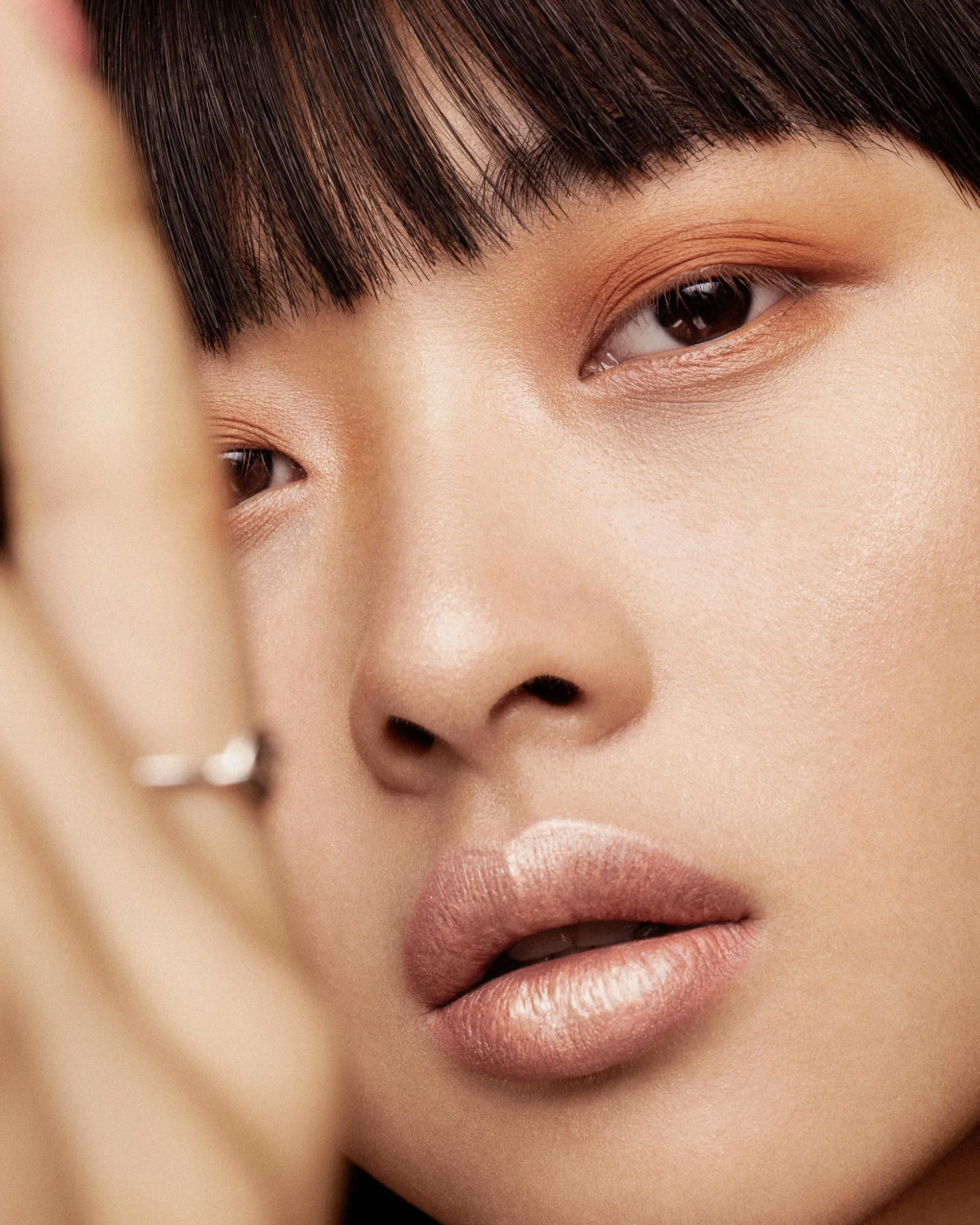 Close-up of a woman's face focusing on her eyes, nose, and lips with makeup, with part of her hand and a ring visible in the foreground.