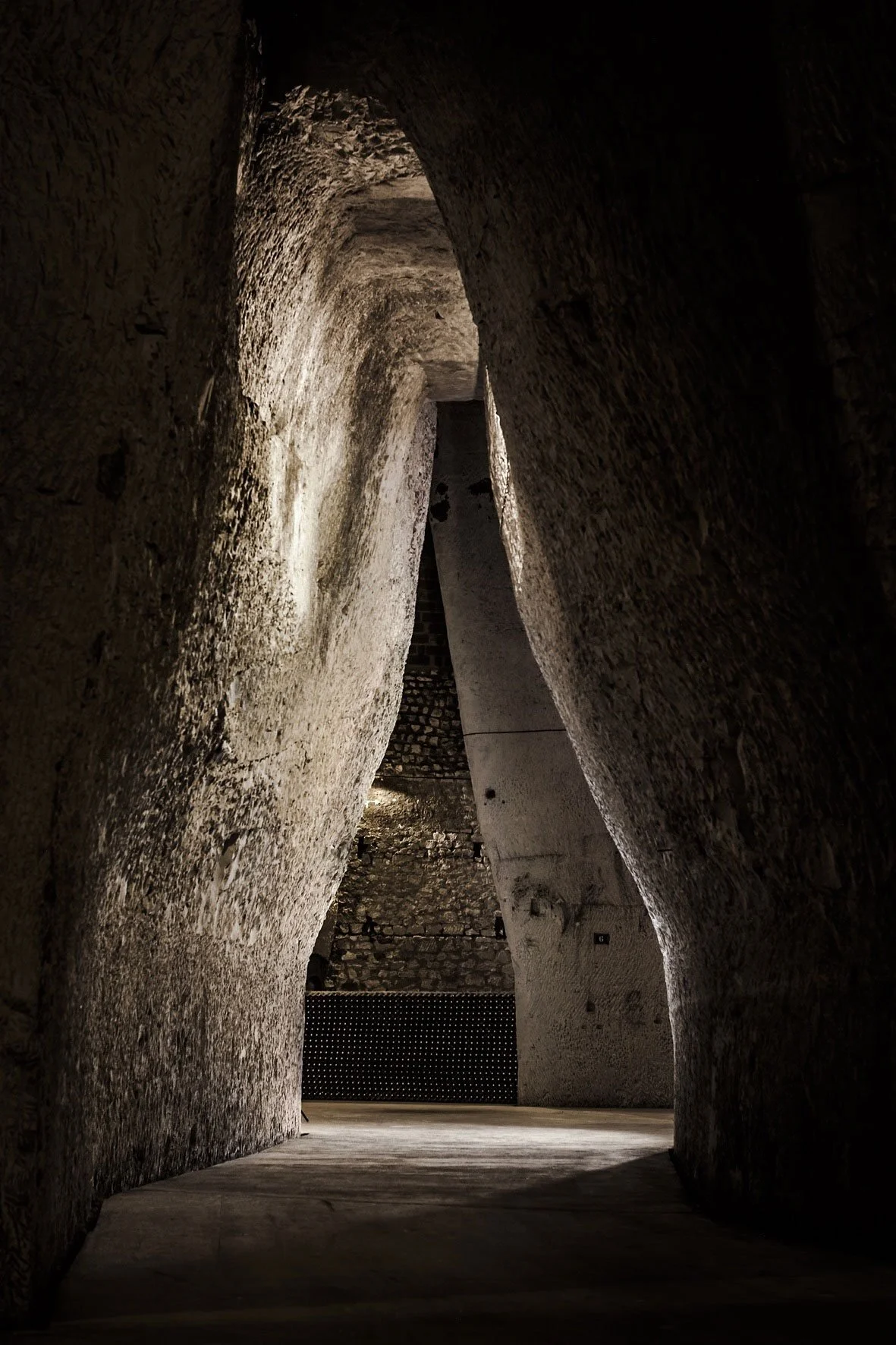 A narrow, dimly lit stone corridor with curved interior walls leading to a bright open space, featuring a modern black railing or panel at the far end.