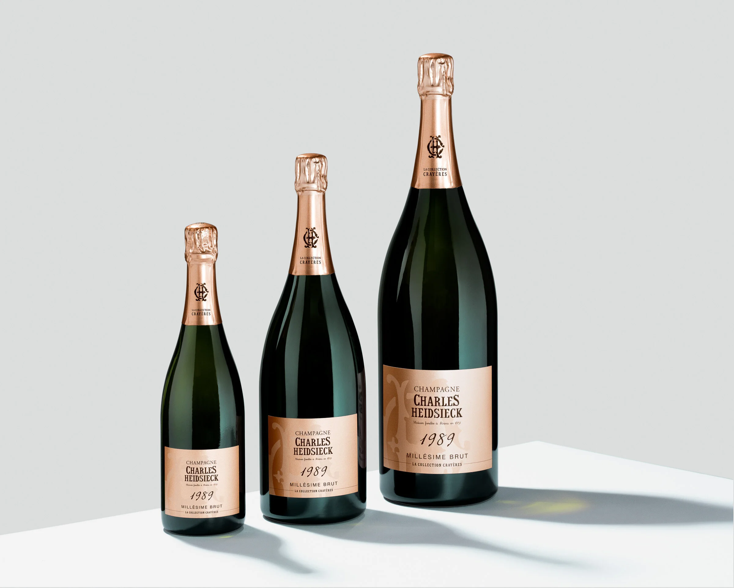 Three bottles of Charles Heidsieck Champagne Millesime Brut 1989, varying in size, placed on a white surface with shadows cast on the right side.