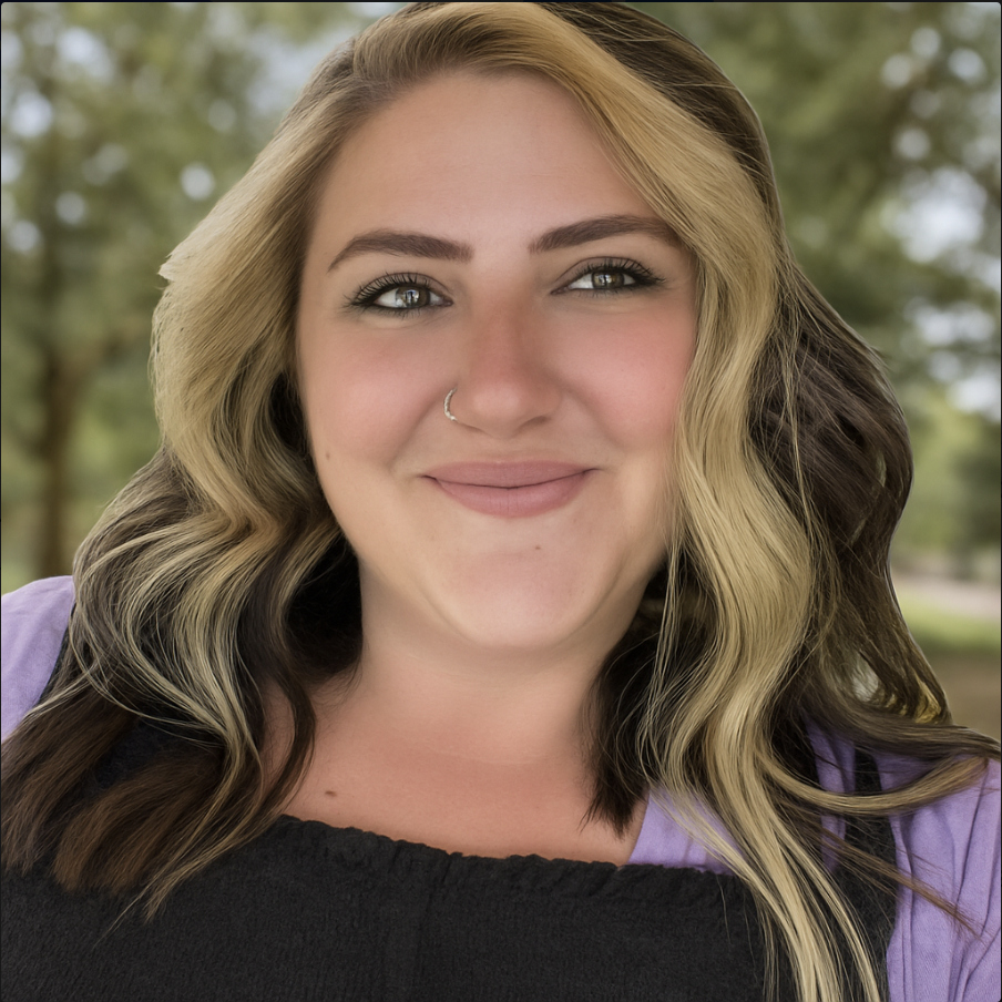 A smiling woman with blonde and brown hair, a nose ring, and wearing a purple top outdoors with blurred trees in the background.
