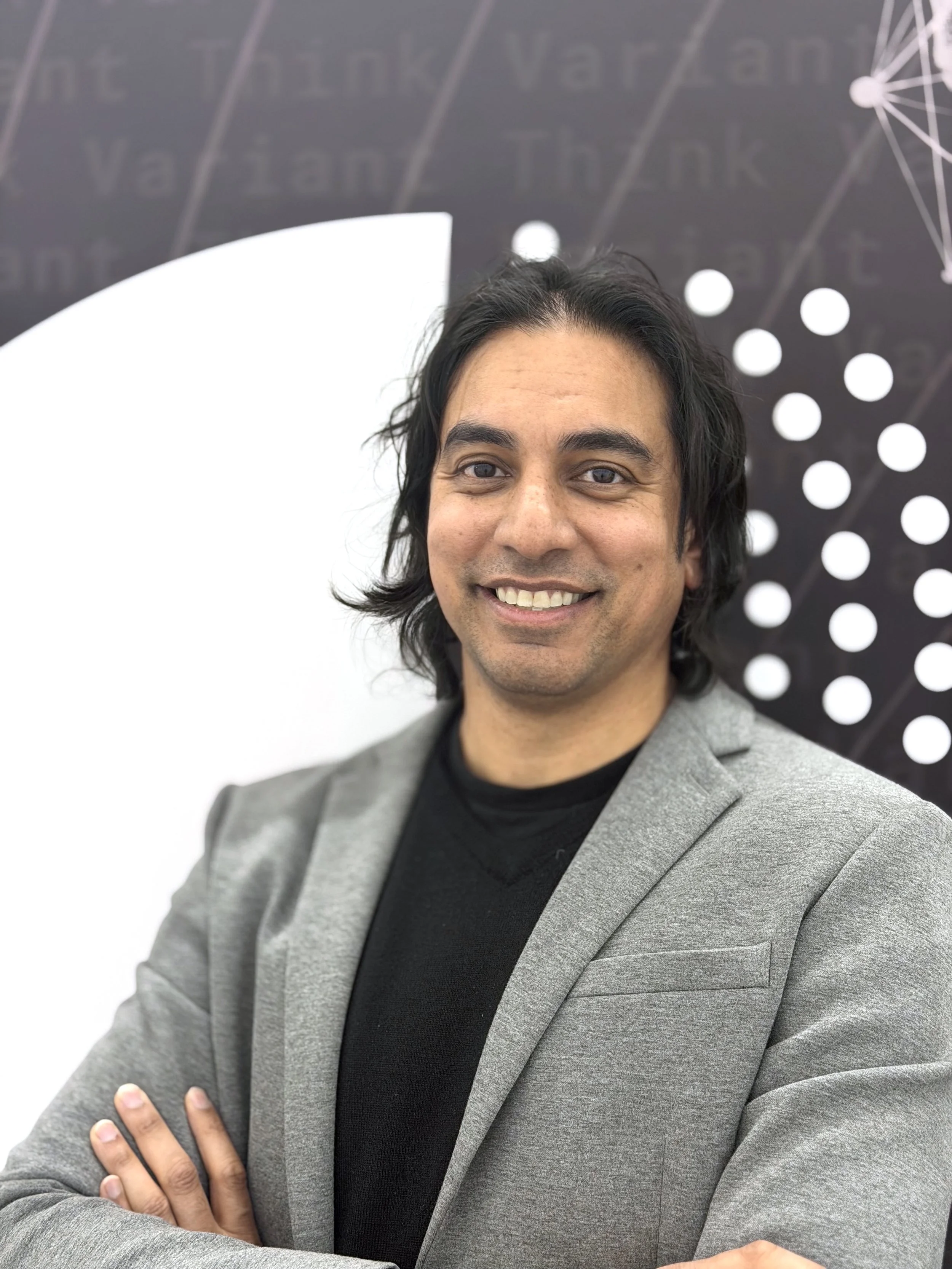A man with shoulder-length black hair, wearing a gray blazer and black shirt, smiling with arms crossed, standing in front of a Think Variant background with white abstract and geometric patterns.