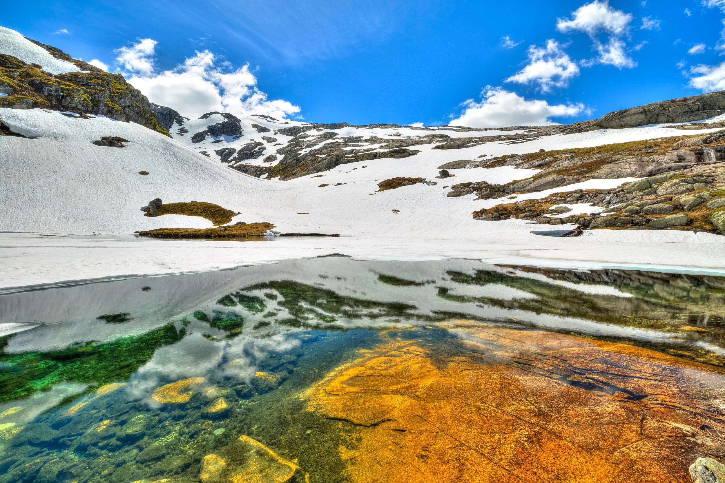 Snø and fjell med en innsjø i forgrunnen, under en blå himmel med noen skyer.