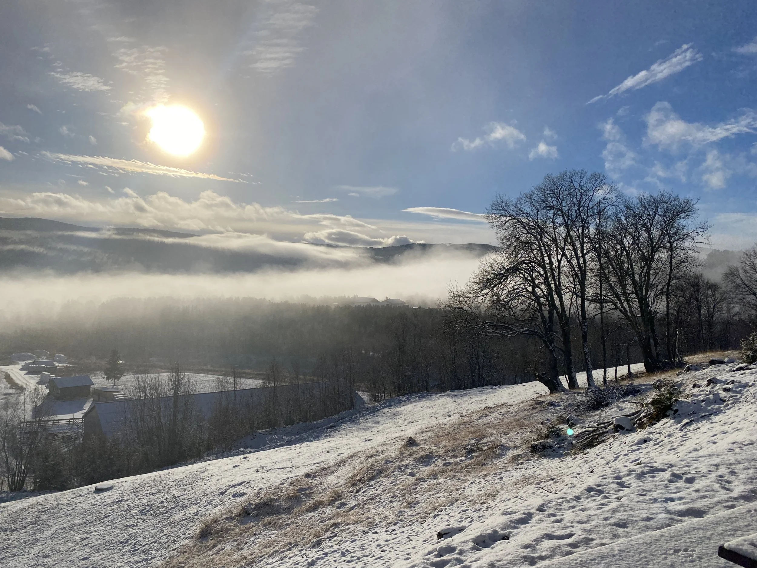 Snødekt ås med bare trær og liten bebyggelse, lavt skydekke og tåke i dalen, solen skinner på himmelen med spredte skyer.
