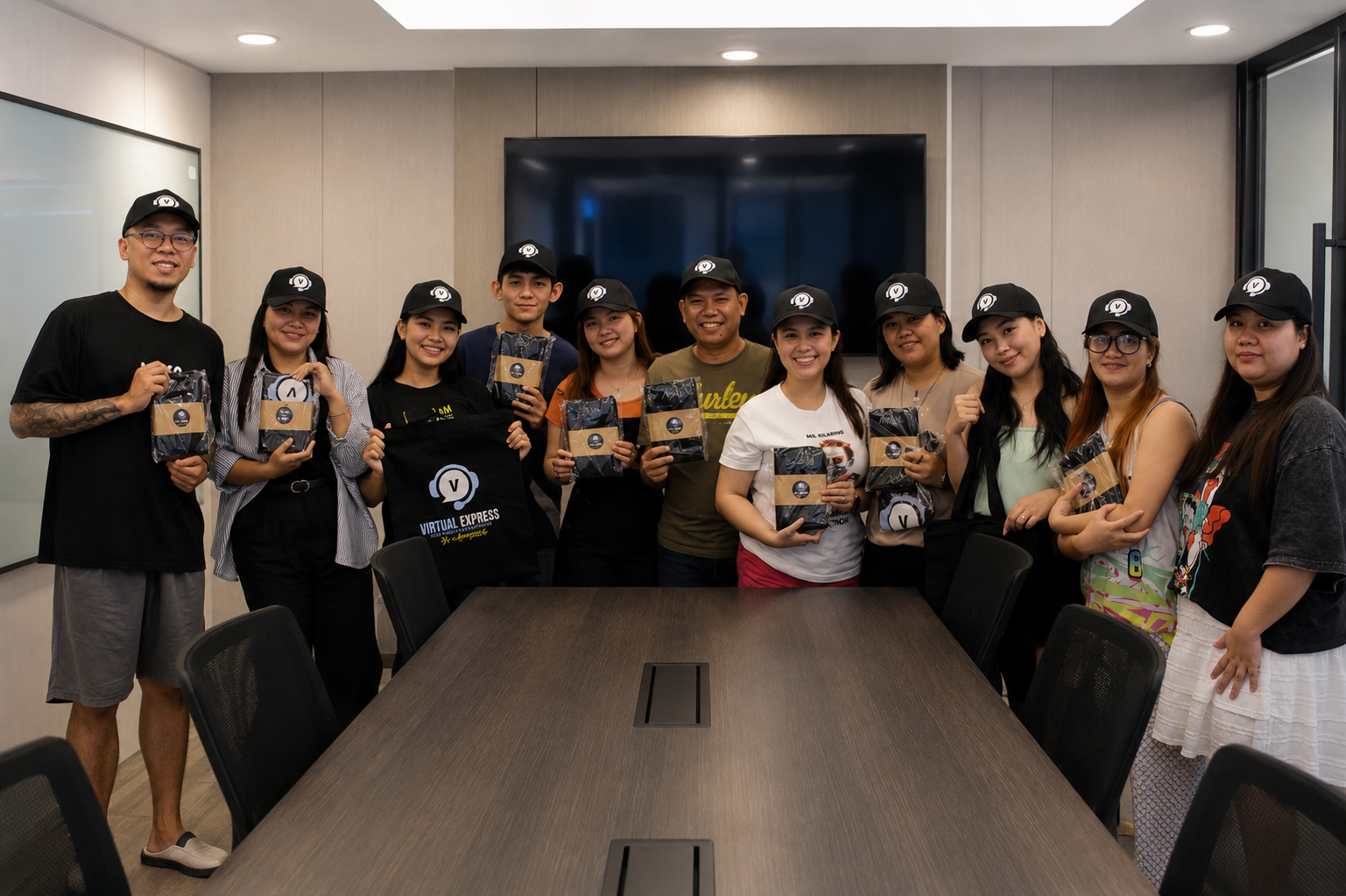 A group of fifteen people, including men and women, standing in a conference room with a large TV screen behind them. They are all smiling and holding packages or bags with the Virtual Express logo, wearing black caps with the same logo, and appear to be celebrating or participating in an event.