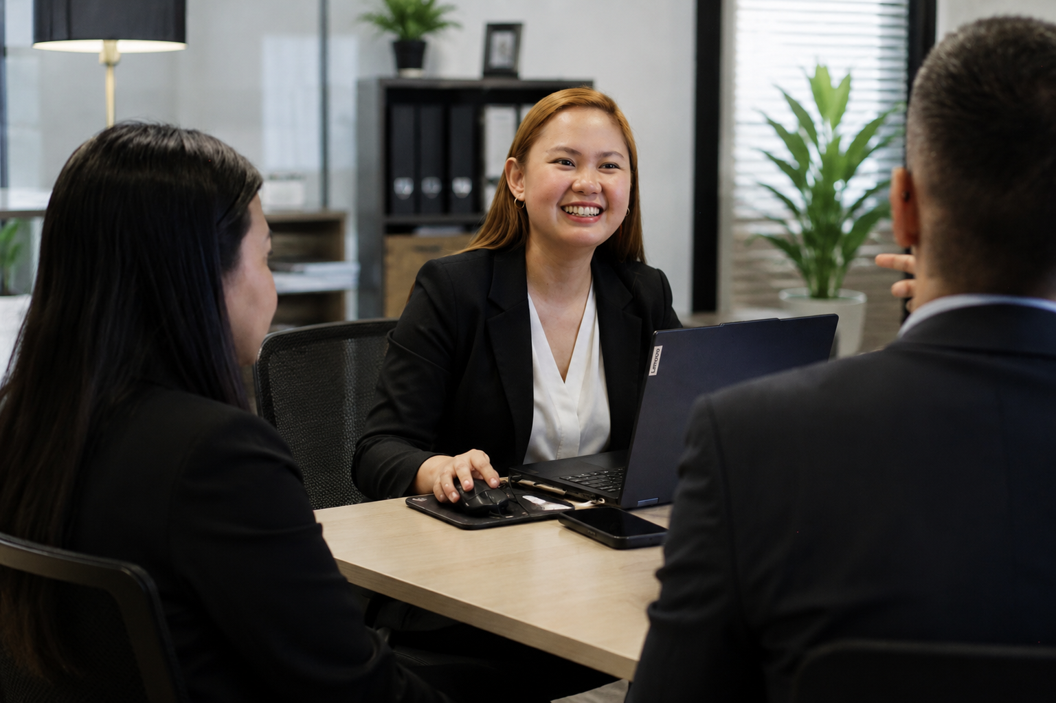 A woman in a black blazer smiling while having a business meeting with two colleagues in an office.