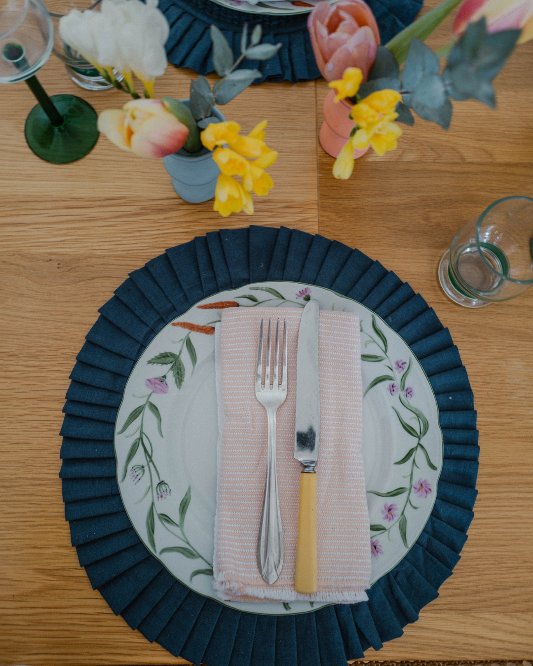 A table setting with a decorative floral plate, a pink-striped napkin, a fork, and a butter knife, surrounded by flower vases and glassware.