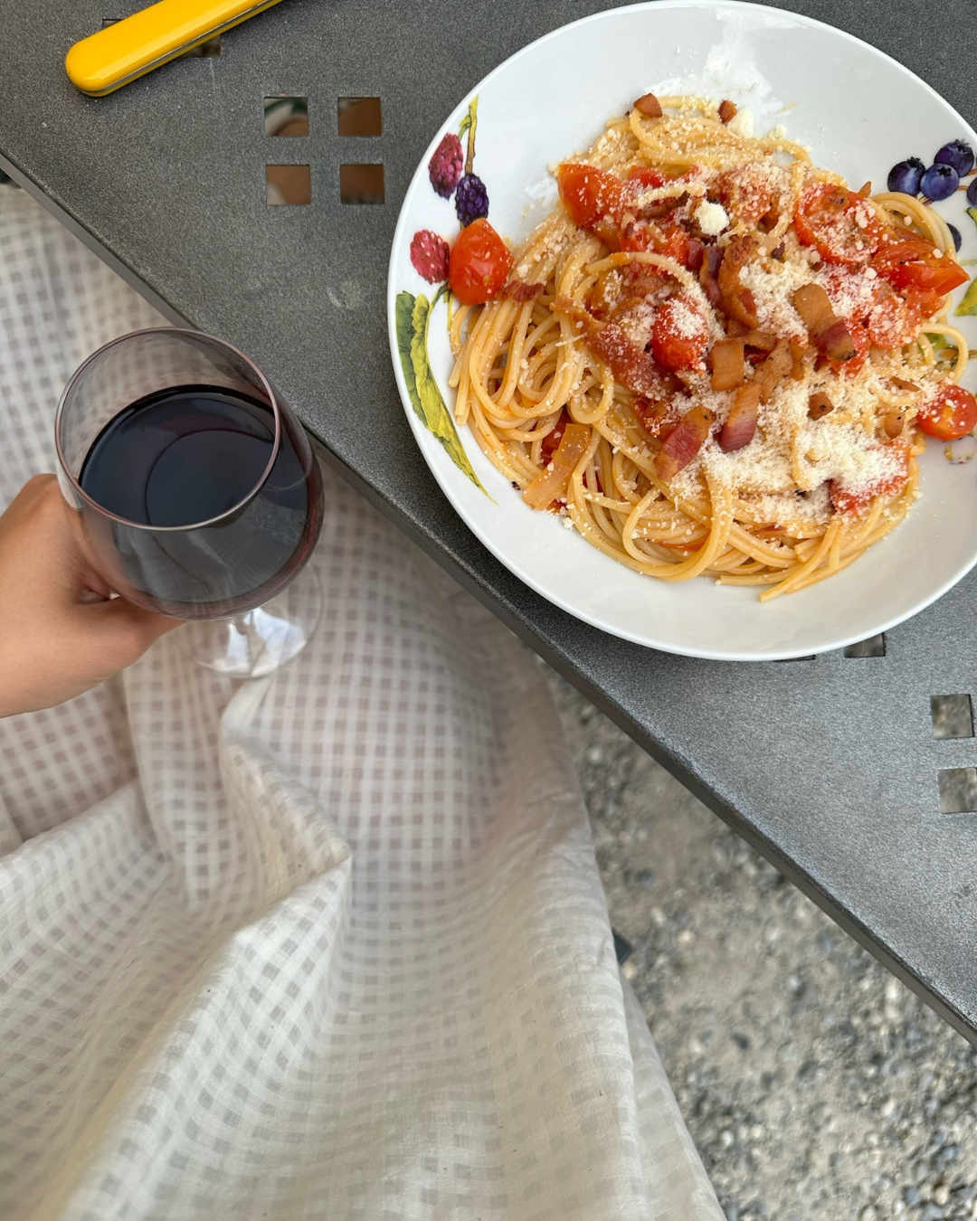 A plate of spaghetti with tomato sauce, grated cheese, and bacon bits, next to a glass of red wine, on a table.
