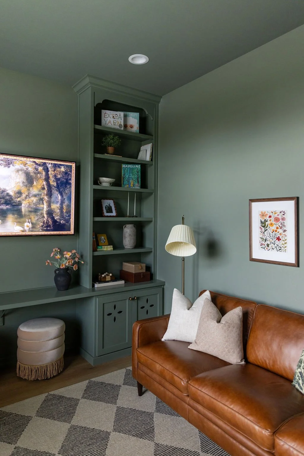Living room with green walls, a brown leather sofa with pillows, a white standing lamp, a framed floral art print, built-in green bookshelves with books and decor, a painting of a lake and swans, a black vase with flowers, and a patterned beige and gray rug.