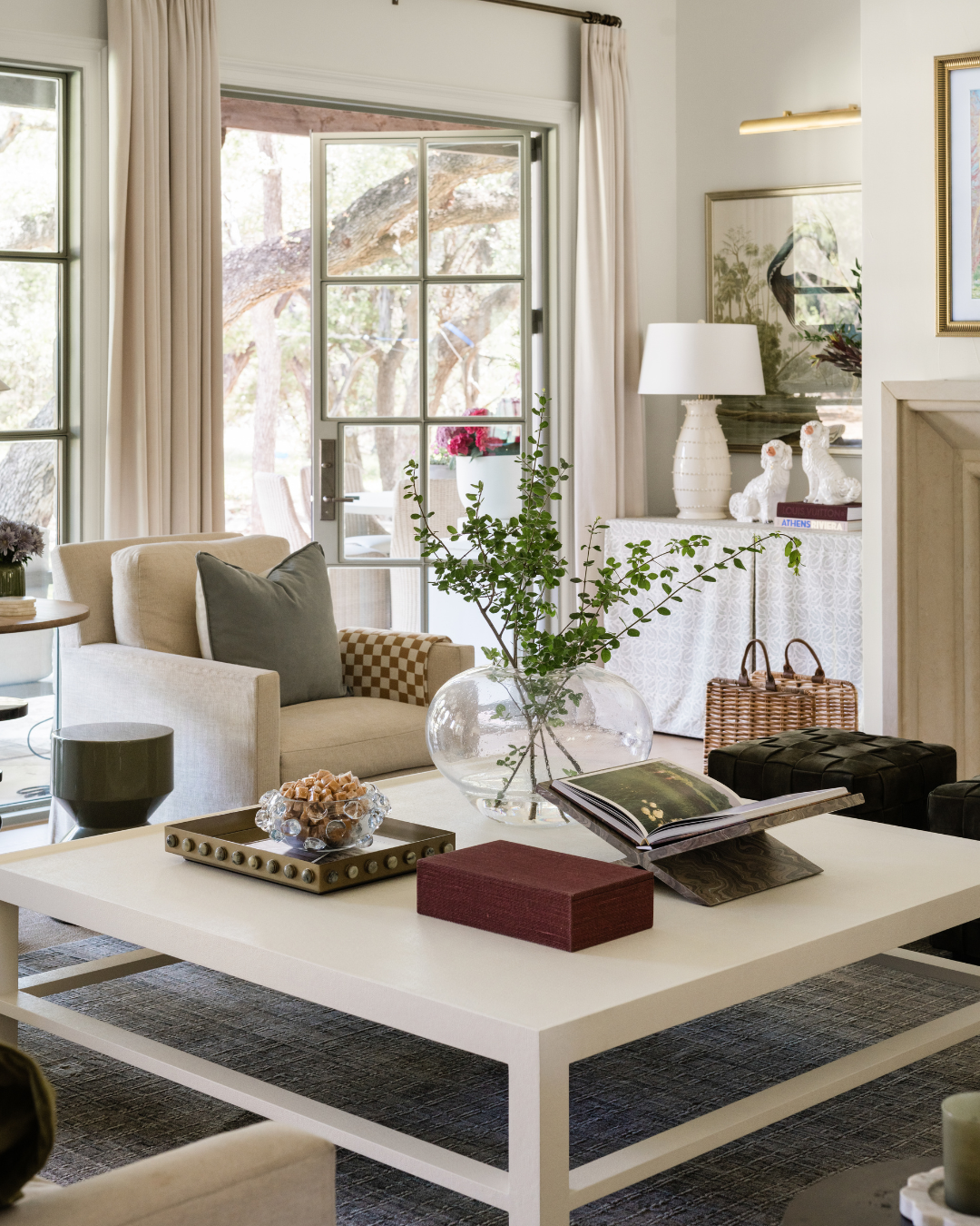 A cozy living room with a white sofa, a coffee table with decorative items, a large glass vase with greenery, a side table, and a sliding glass door opening to an outdoor patio with trees.
