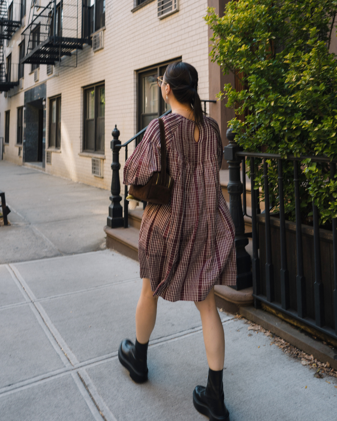 A woman with dark hair walking on a city sidewalk with brick residential buildings and green bushes on the side.