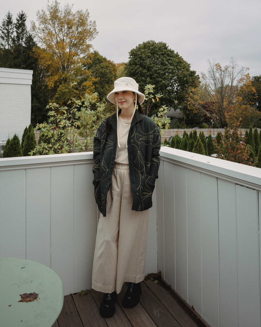 A woman standing on a wooden porch with white railing, wearing a beige bucket hat, cream top, black quilted jacket with gold stitching, beige wide-leg pants, and black boots, with a backyard with trees and bushes in fall colors in the background.