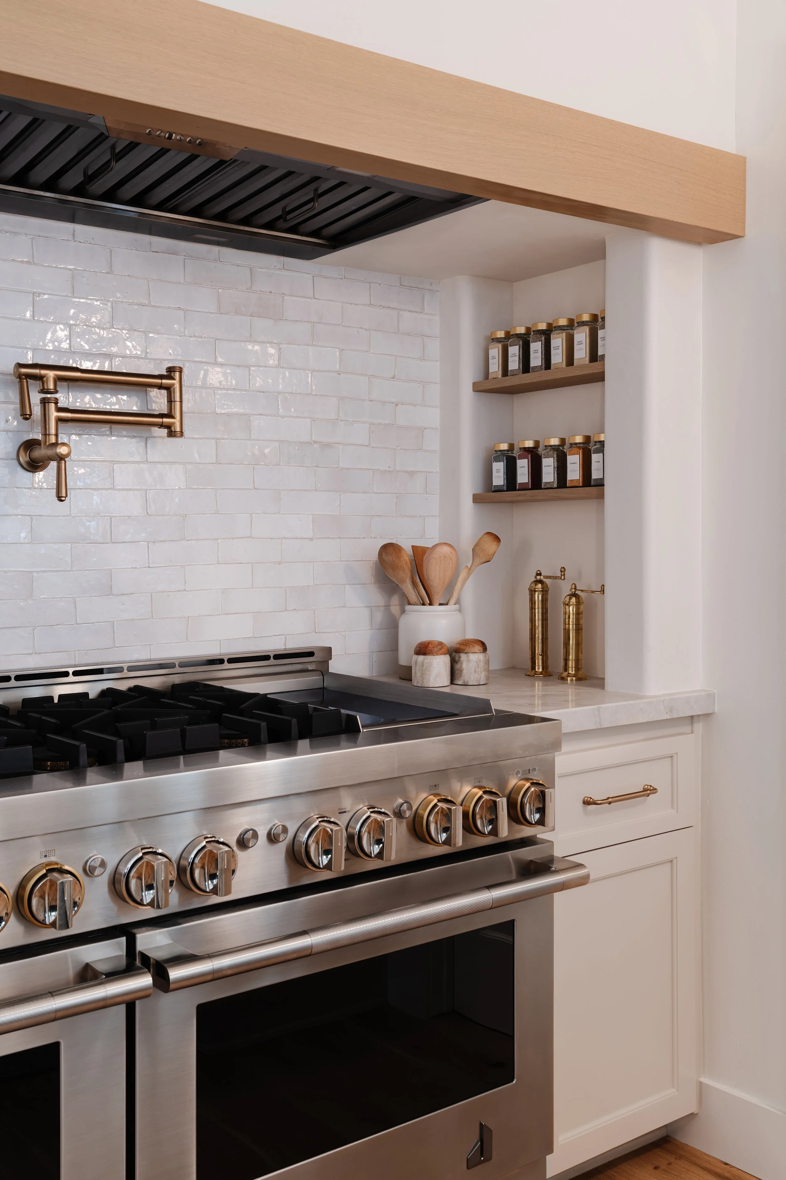 Modern kitchen with stainless steel gas stove, white cabinetry, wooden shelves with spice jars, white brick backsplash, and gold kitchen accessories.