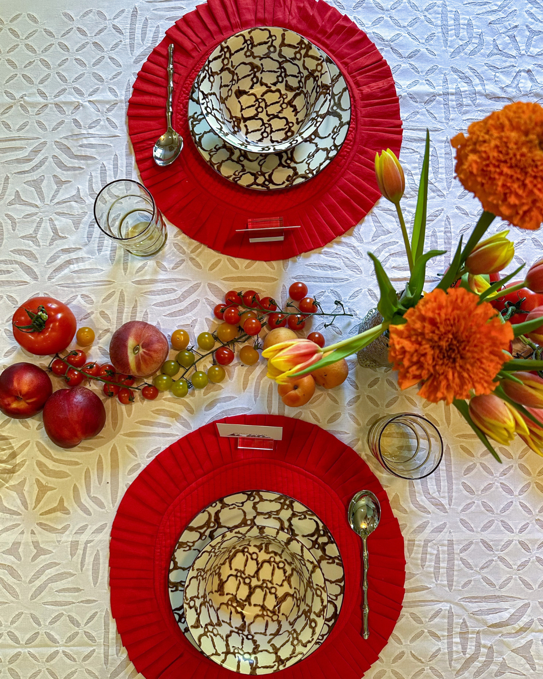 Top view of a table set for two with red placemats, decorative black and white patterned bowls, and clear glasses, decorated with a floral centerpiece of lilies and marigolds, along with strawberries, apples, and cherry tomatoes.
