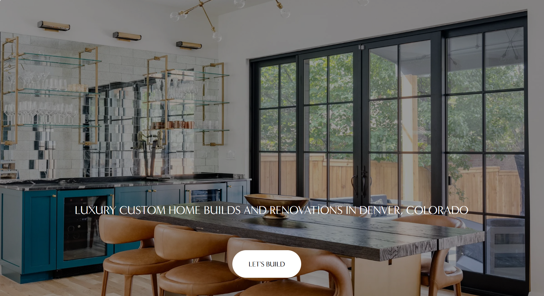 Modern kitchen with blue cabinets, glass shelves with glassware, a black countertop, and large black-framed glass doors leading to an outdoor patio with trees and a wooden fence.