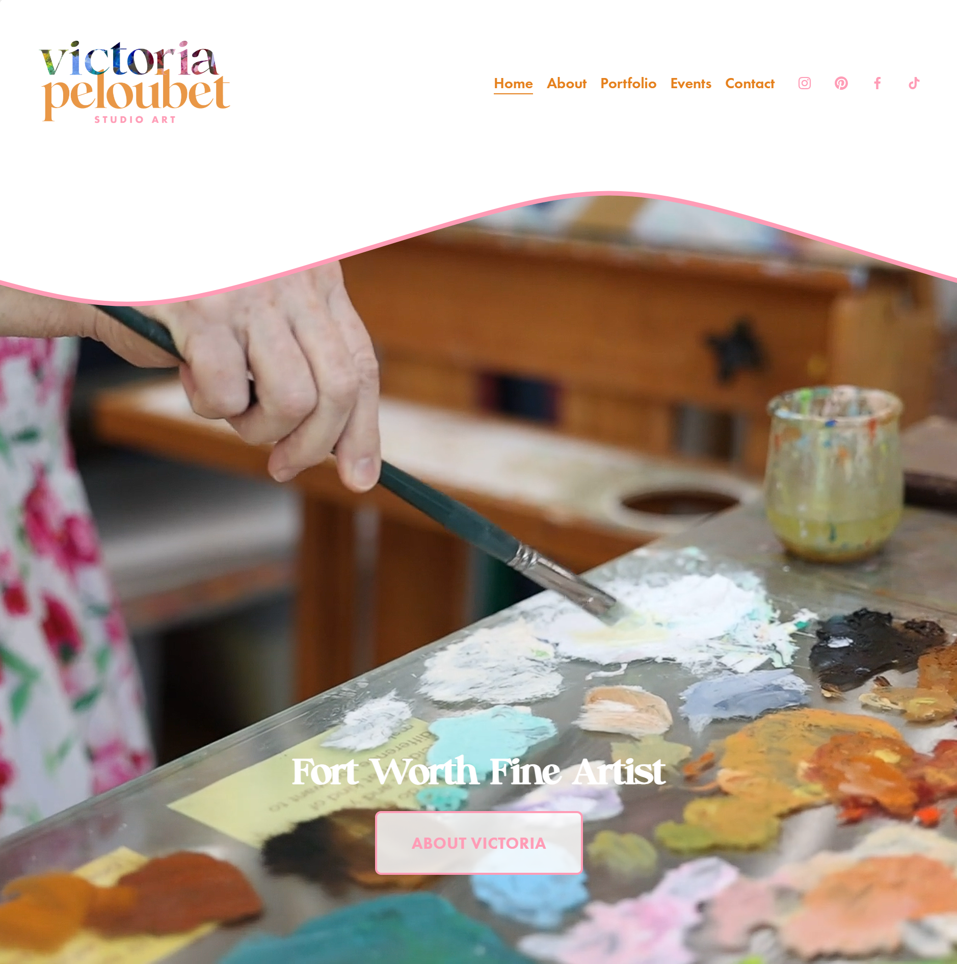 A person painting at an art studio table with colorful paints and a palette, with a wooden art table in the background. The person is not fully visible, only their hand holding a paintbrush.