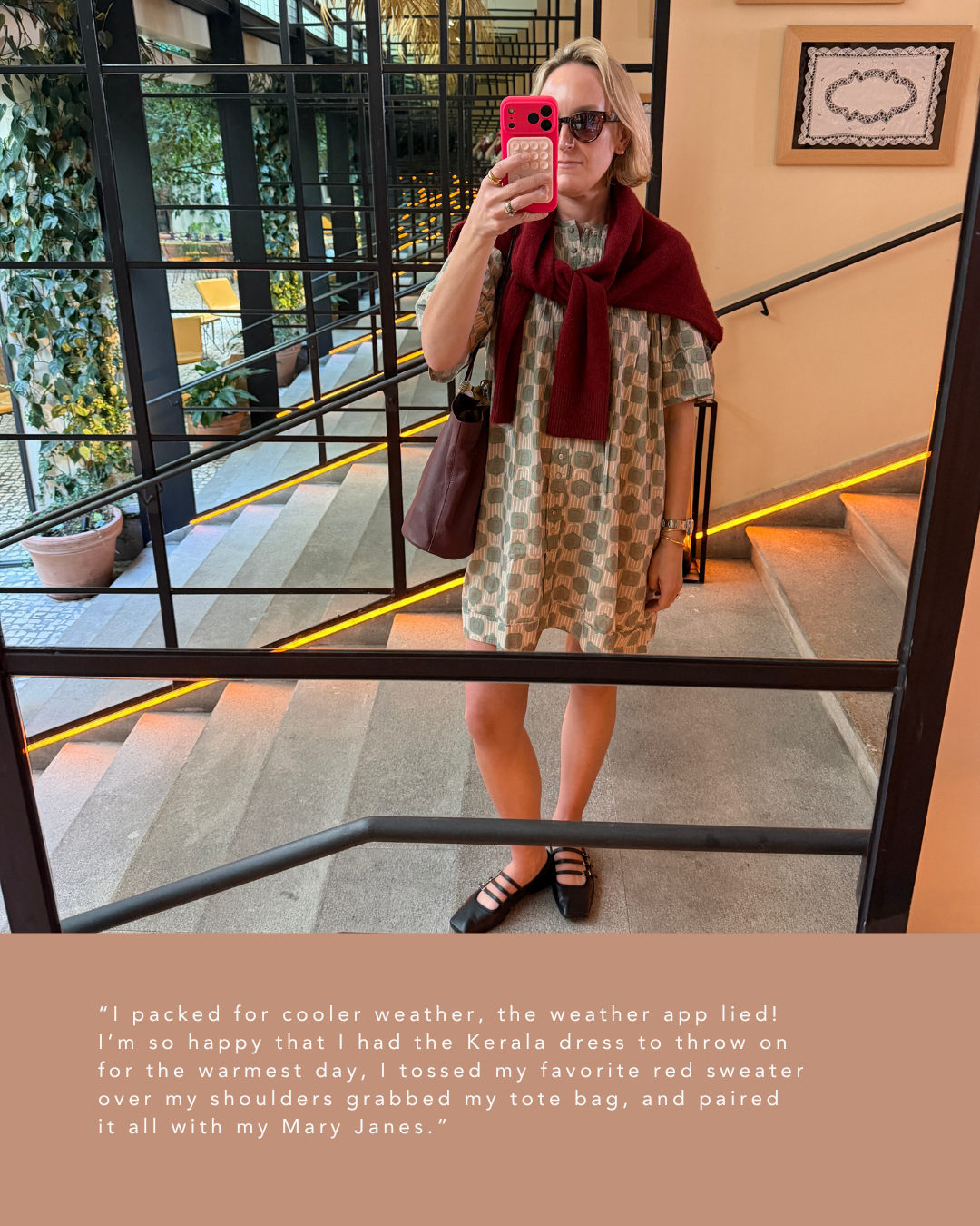 A woman taking a selfie in a mirror, wearing sunglasses, a patterned dress, a red sweater draped over her shoulders, black shoes, and carrying a brown tote bag, standing on a staircase in a decoratively lit interior.