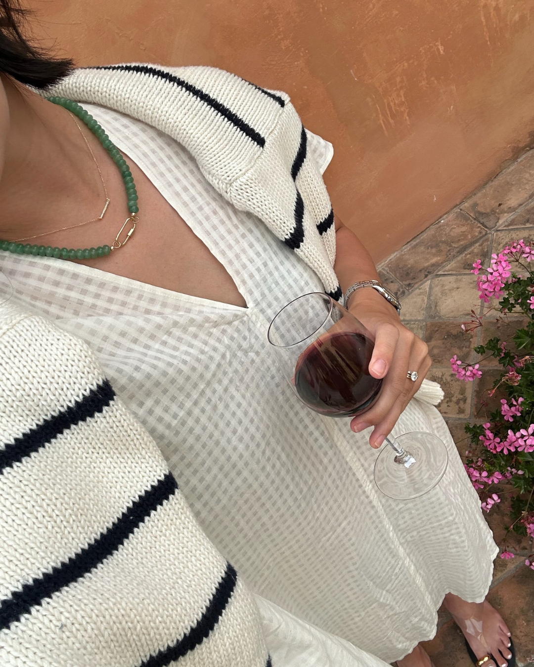 Woman wearing a white gingham dress, a knitted cardigan with black stripes, green bead necklace, and gold jewelry, holding a glass of red wine, standing near a pink flowering plant.