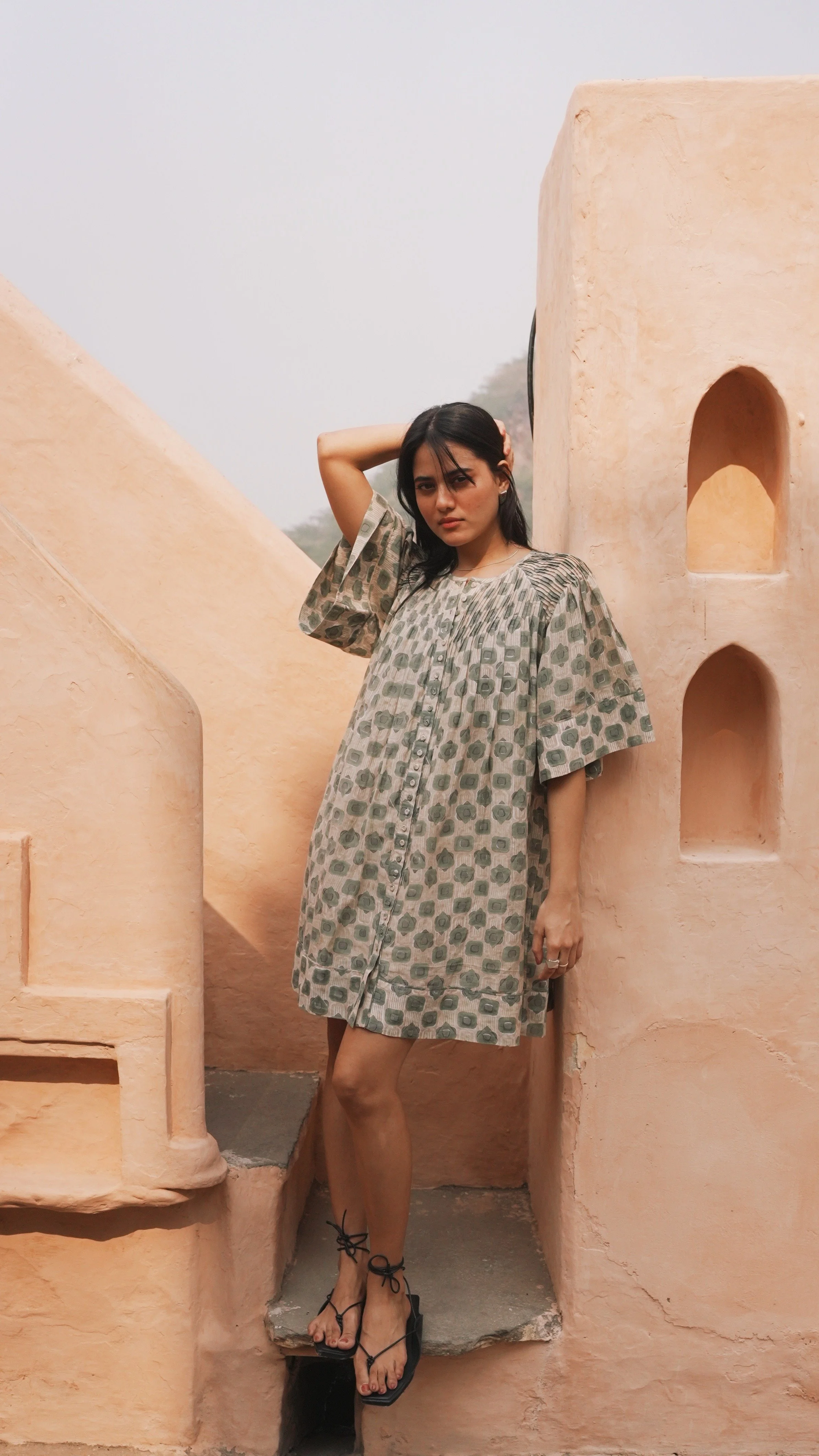 A woman in a patterned dress standing next to a textured peach-colored wall with arched openings, outdoors on a foggy day.