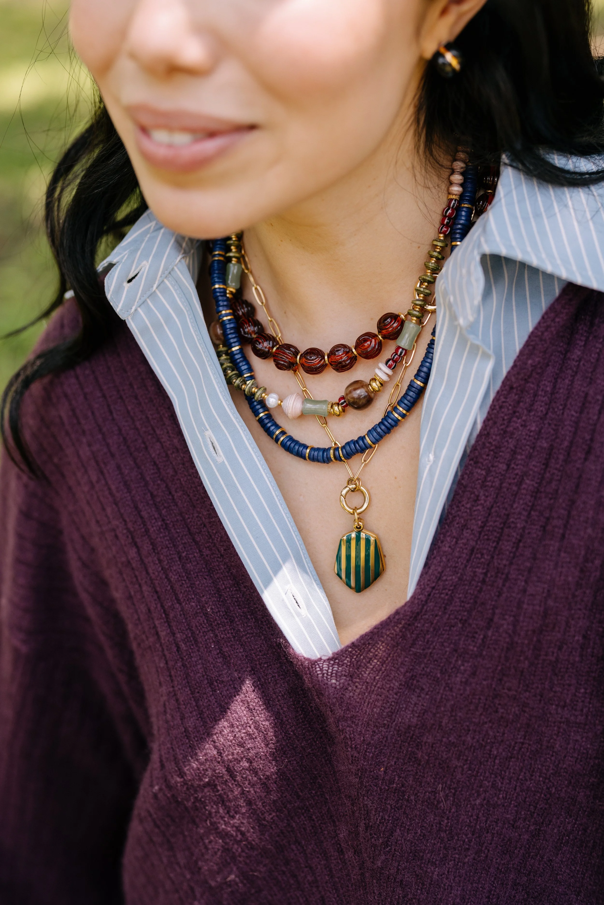 Close-up of a woman wearing layered statement necklaces, a striped collared shirt, and a purple sweater, with a grassy outdoor background.