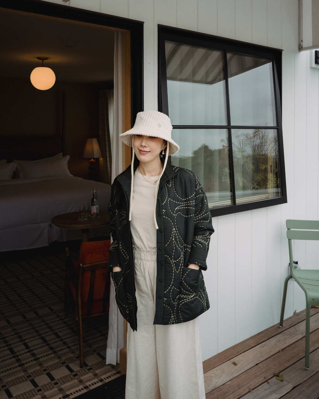 A woman standing outside a building with a large window. She is wearing a white bucket hat, a black quilted jacket with gold stitching, and cream-colored wide-leg pants. She is smiling slightly with her eyes closed.