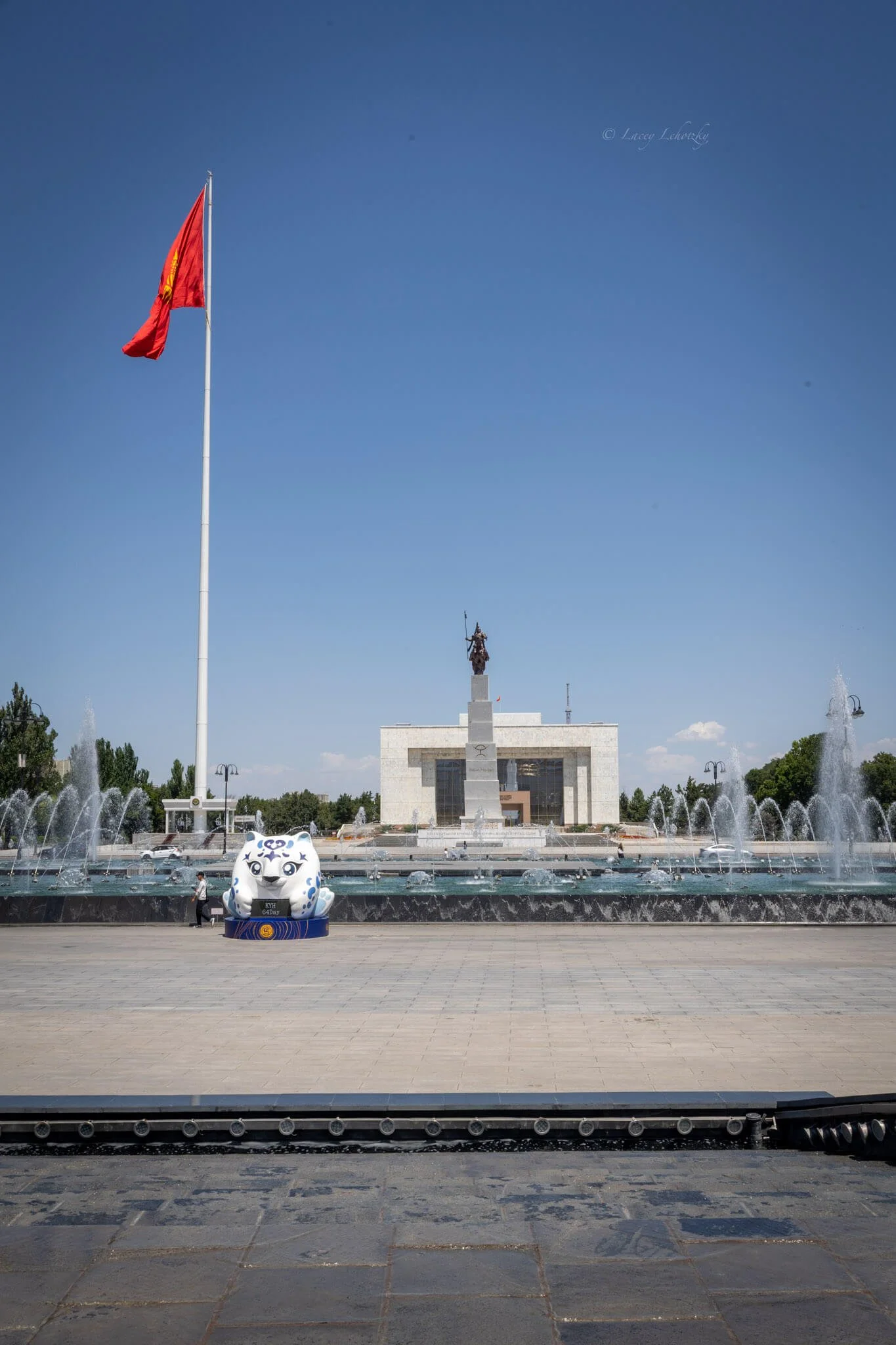a bronze statue of a man riding a horse, a massive red kyrgyz flag, and marable buildings line the ala too square bishkek kyrgyzstan