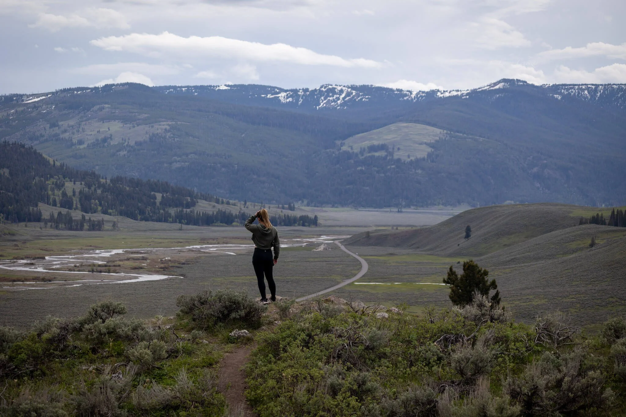 a woman stands on the edge of a cliff overlooking a sweeping valley with snow capped mountains in the distance in yellowstone national park
