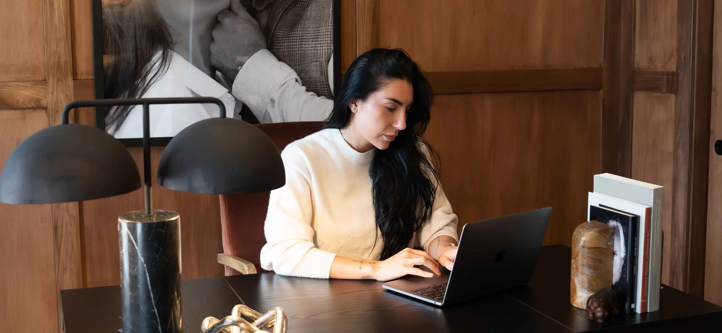A woman with black hair working on a laptop at a dark wooden desk in a room with wooden walls, with decorative books and objects on the desk.