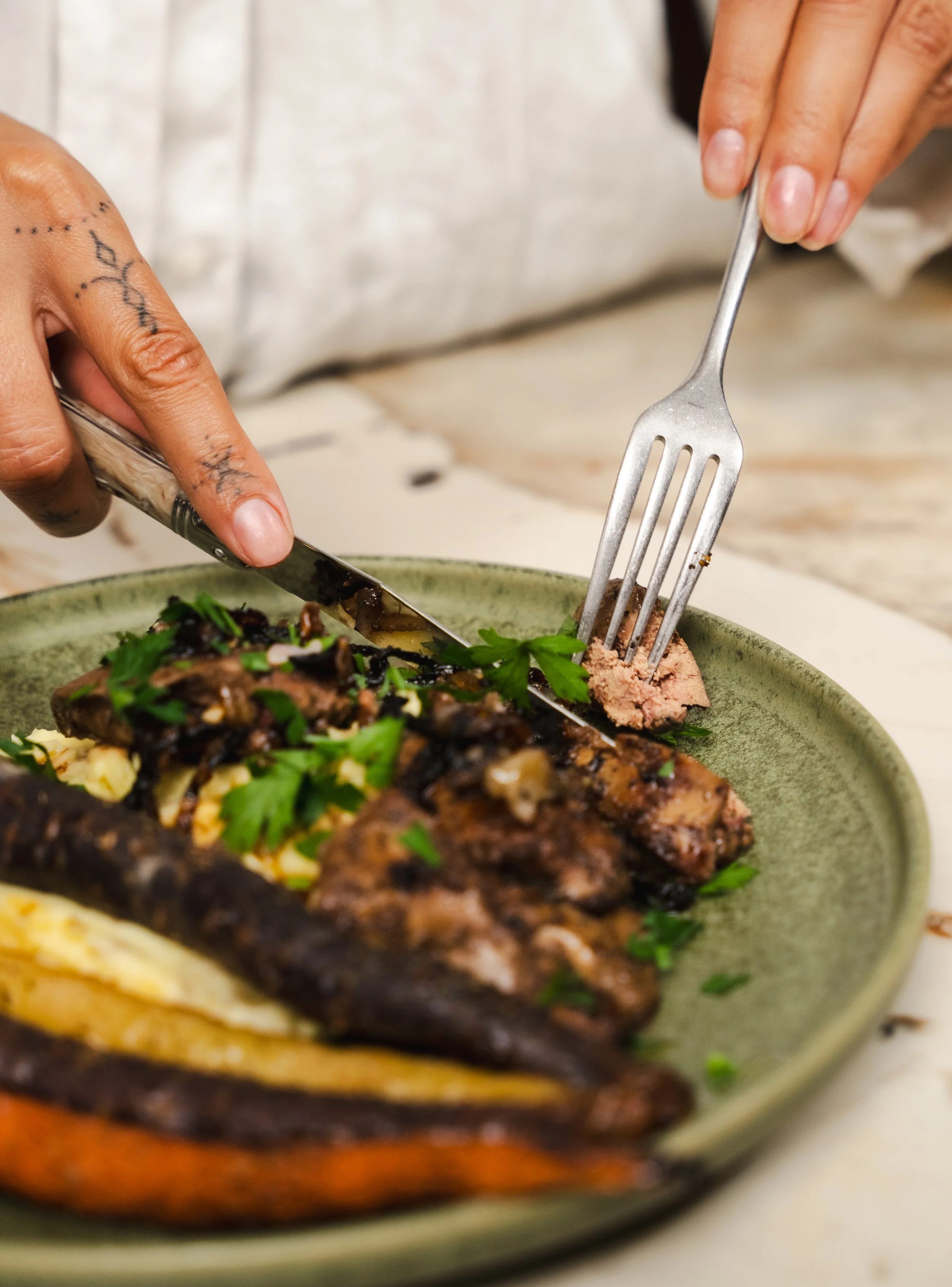 A person is serving sliced meat from a green bowl with a fork and knife. The person's hands have tattoos and a white long sleeve shirt is visible. The dish has green herbs on top.