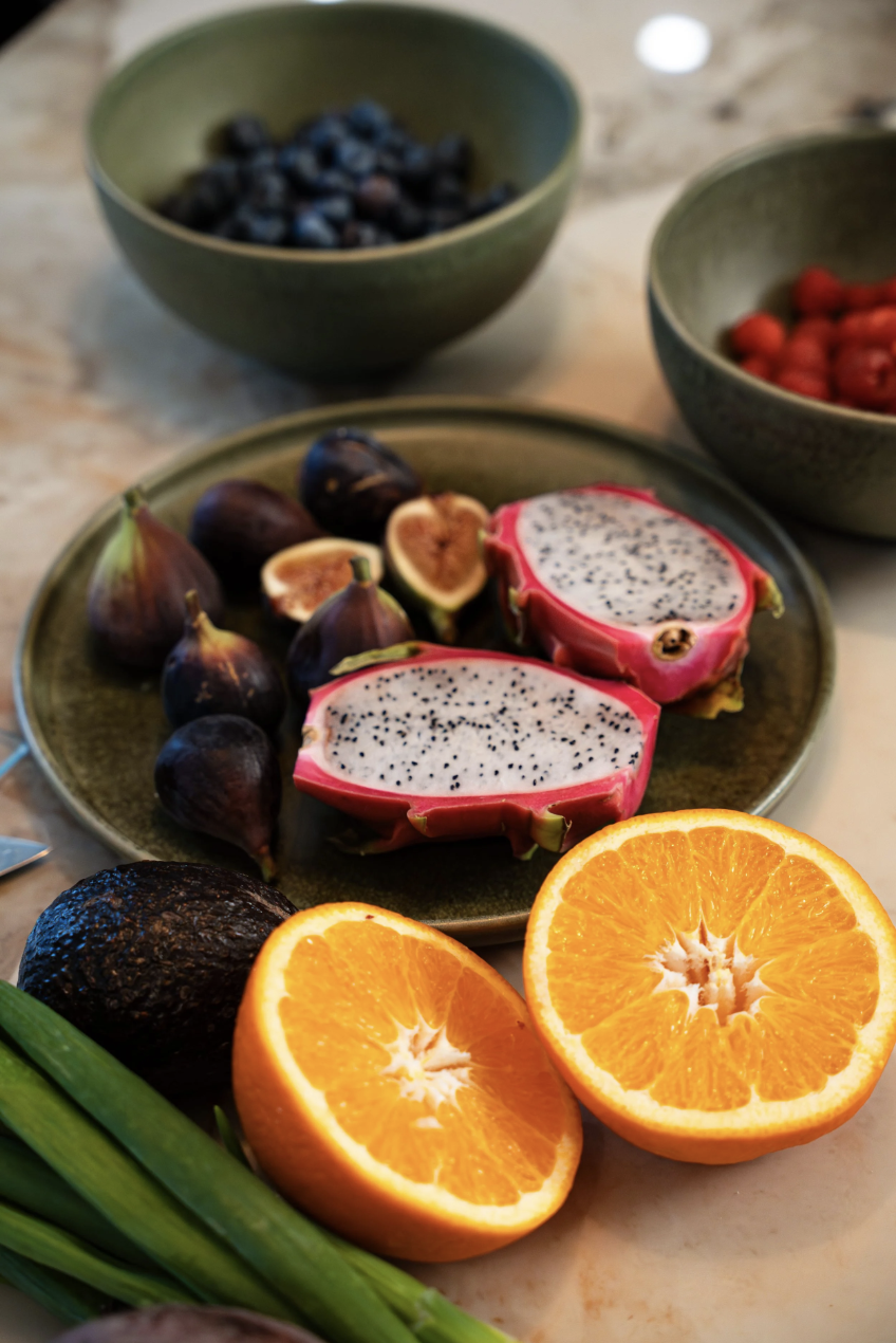 Assorted fresh fruits including halved oranges, dragon fruit, figs, figs in a green bowl, blueberries, strawberries, and an avocado, arranged on a light-colored surface.