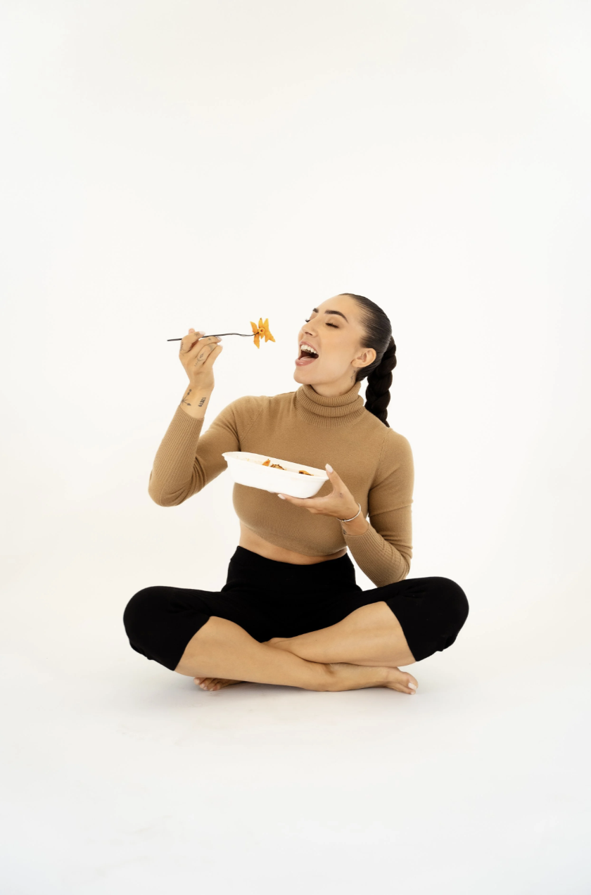 A young woman sitting cross-legged on the floor, holding a white bowl of food in one hand and a fork in the other, smiling and about to eat pasta.