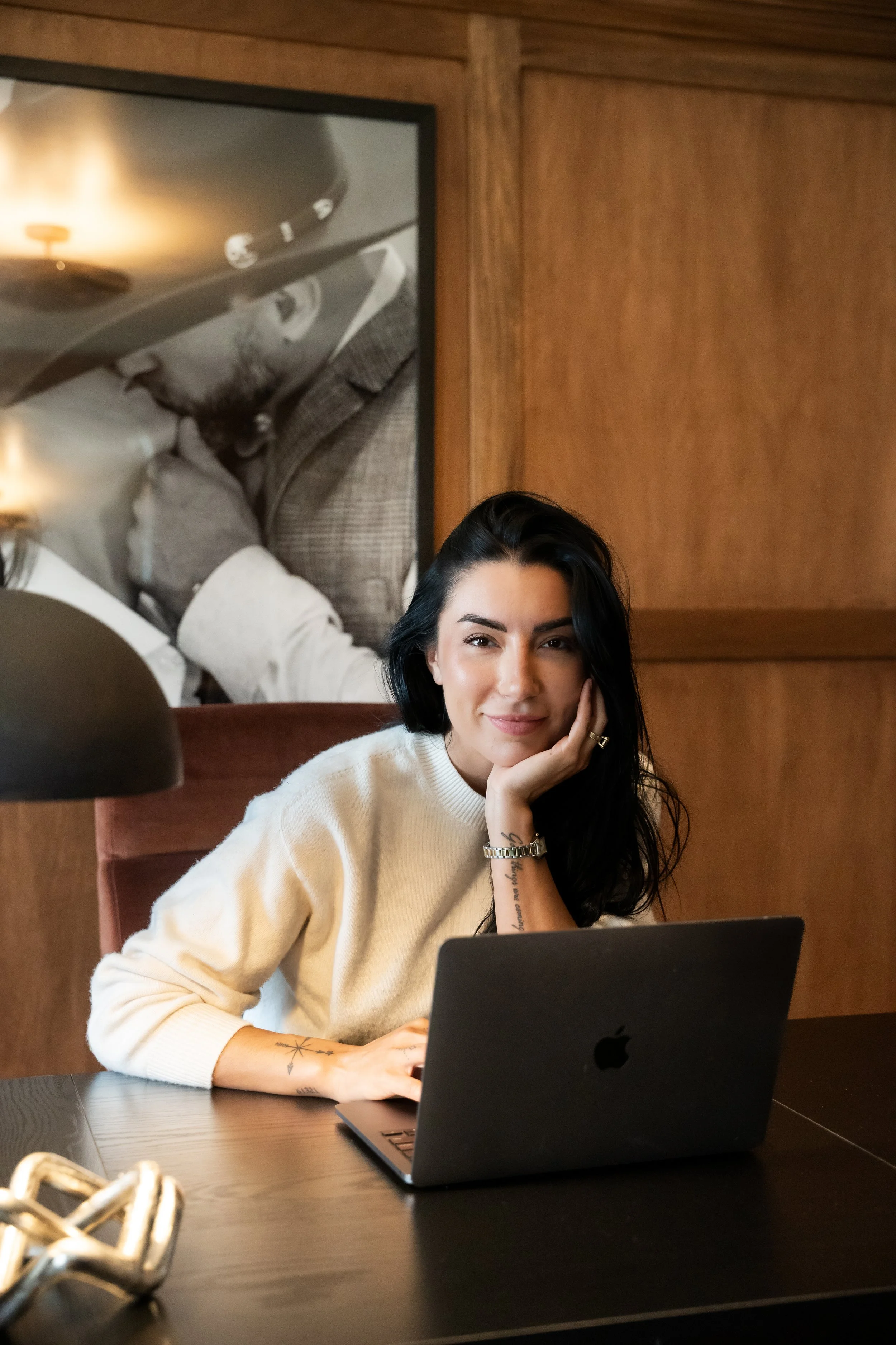 A woman with black hair and tattoos on her arm smiling and resting her face on her hand while sitting at a table with a laptop in front of her.