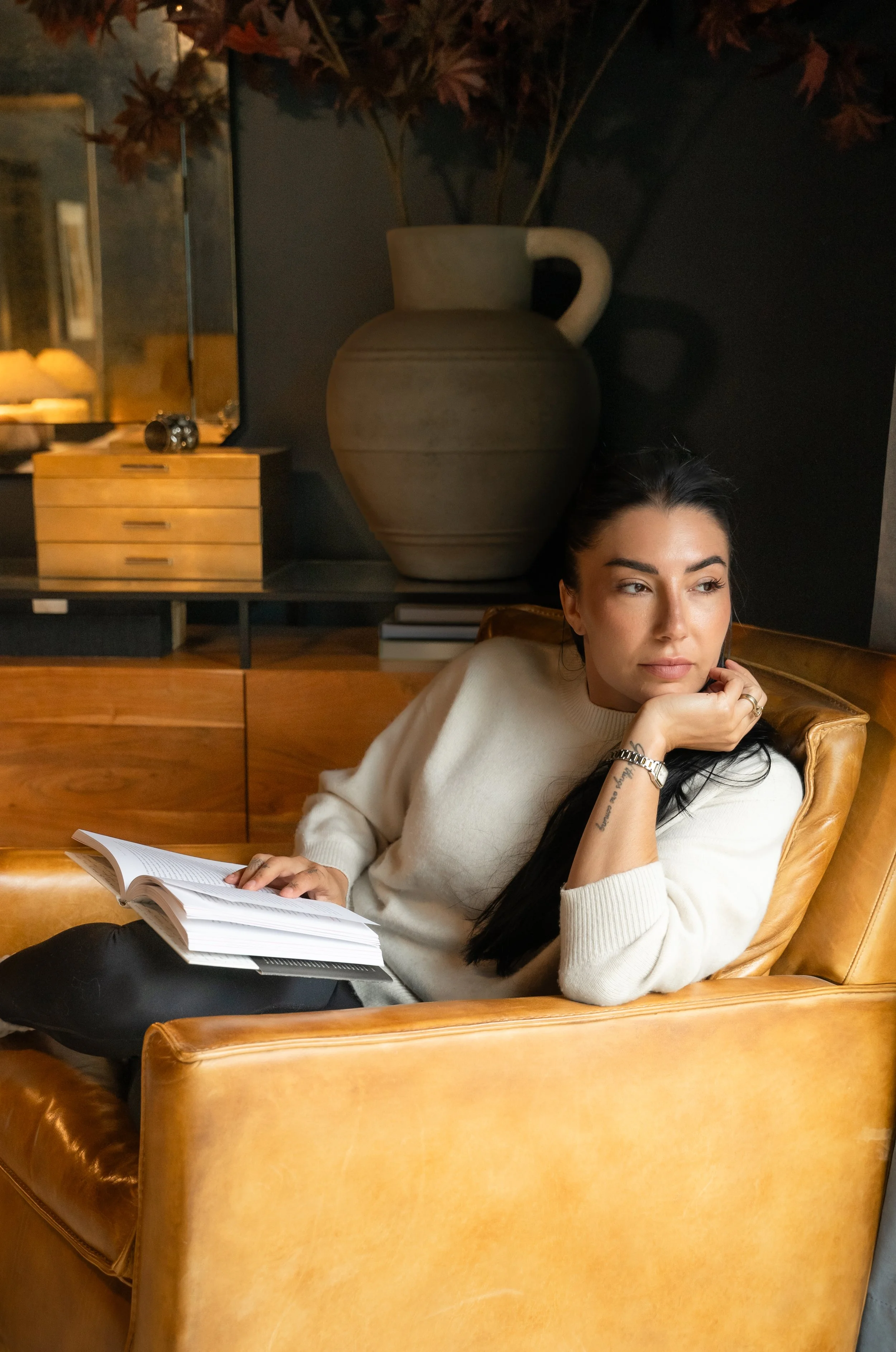 A woman with long dark hair and light skin, wearing a cream-colored sweater and black pants, is sitting on a tan leather chair in a cozy indoor setting. She is reading a book and resting her chin on her hand.