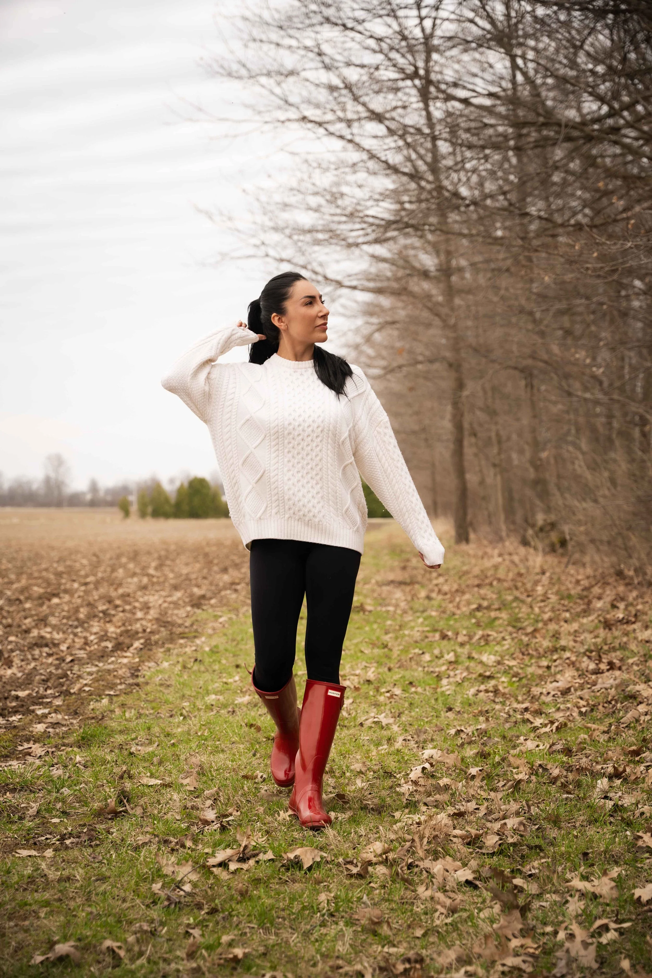A woman with black hair, wearing a white sweater, black leggings, and red rain boots, is walking outdoors on a path covered with fallen leaves, with bare trees in the background during late fall or early winter.