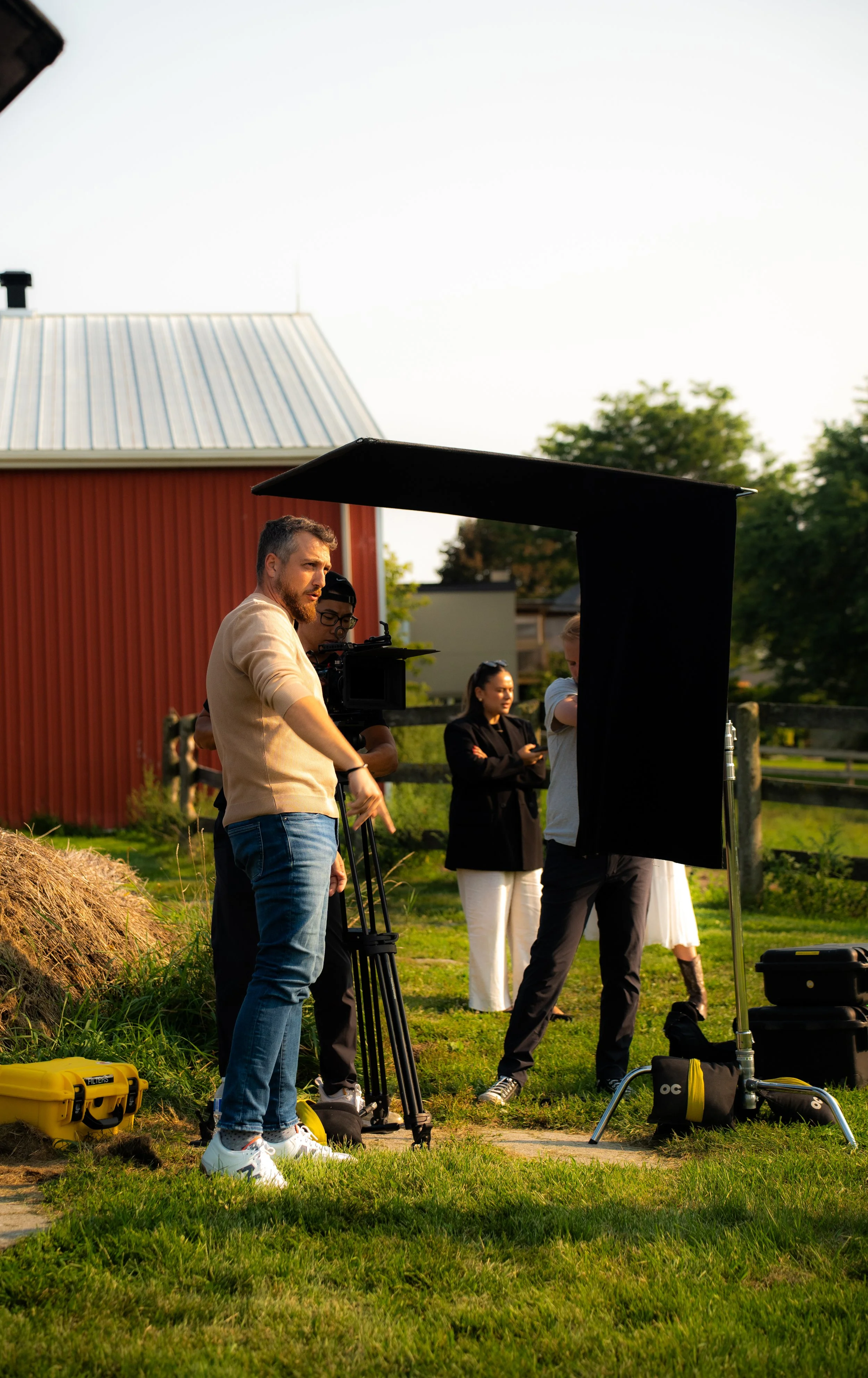People filming outdoors near a red barn with equipment, including a camera on a tripod, lighting, and cases, during early evening.