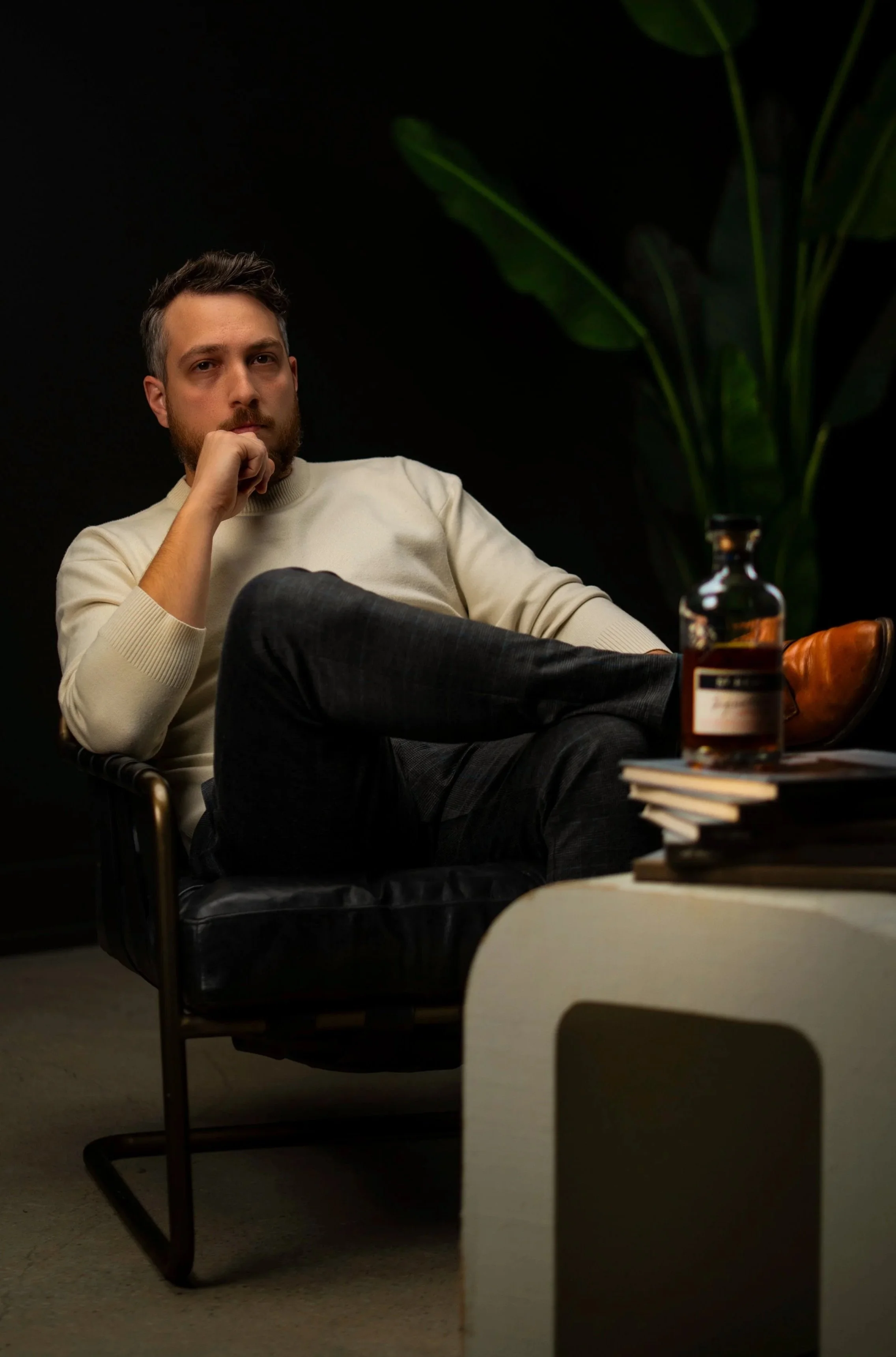 A man sitting in a black leather chair with one leg crossed over the other, wearing a cream sweater and dark pants, in a dimly lit room with dark background and potted plants, with books and a bottle on a table nearby.