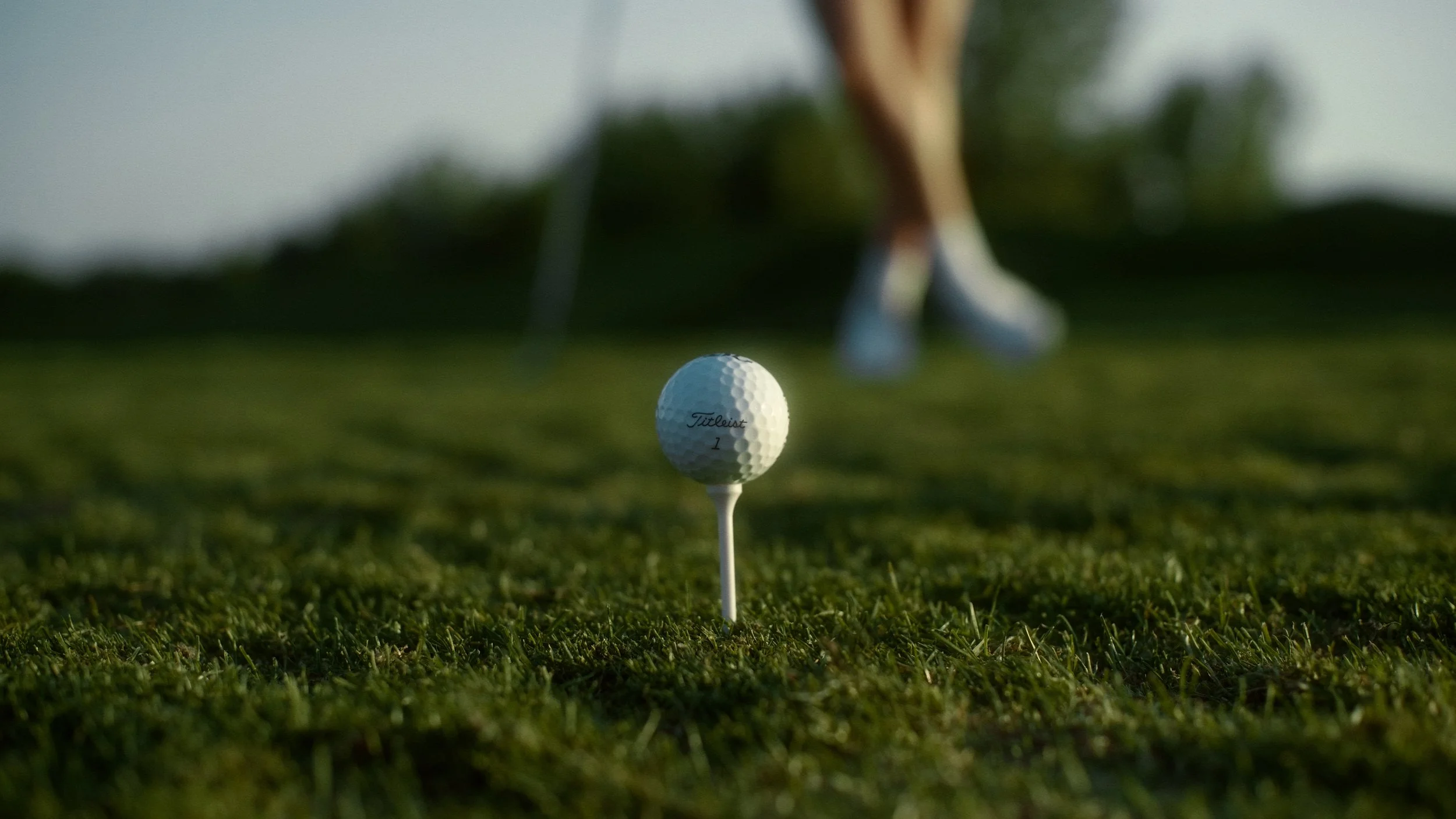 A golf tee with a Titleist golf ball on top is on a grassy golf course. In the background, blurred, is a person's legs walking.
