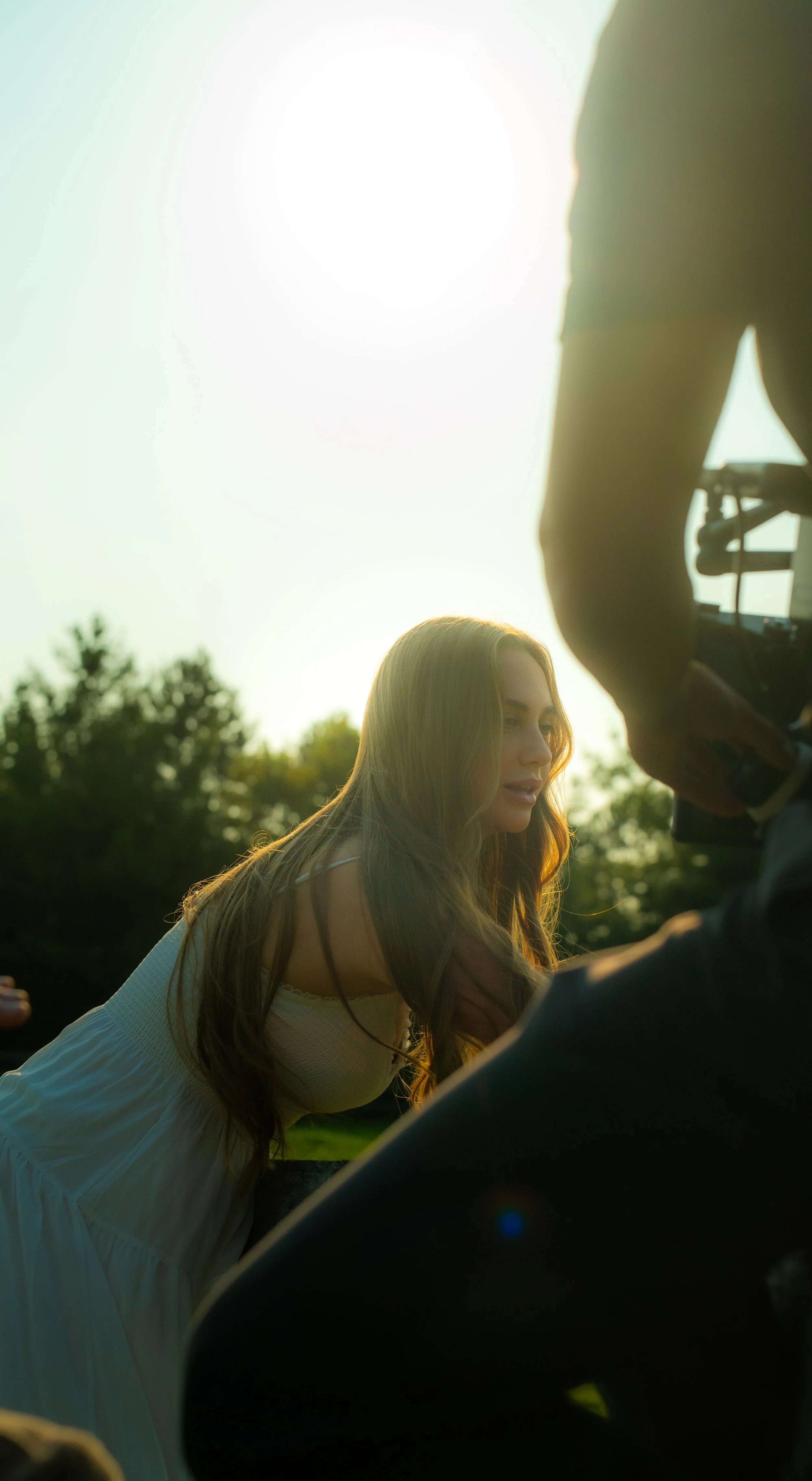 A woman with long, wavy brown hair wearing a white dress, leaning forward outdoors during sunset, with trees and a bright sky in the background.