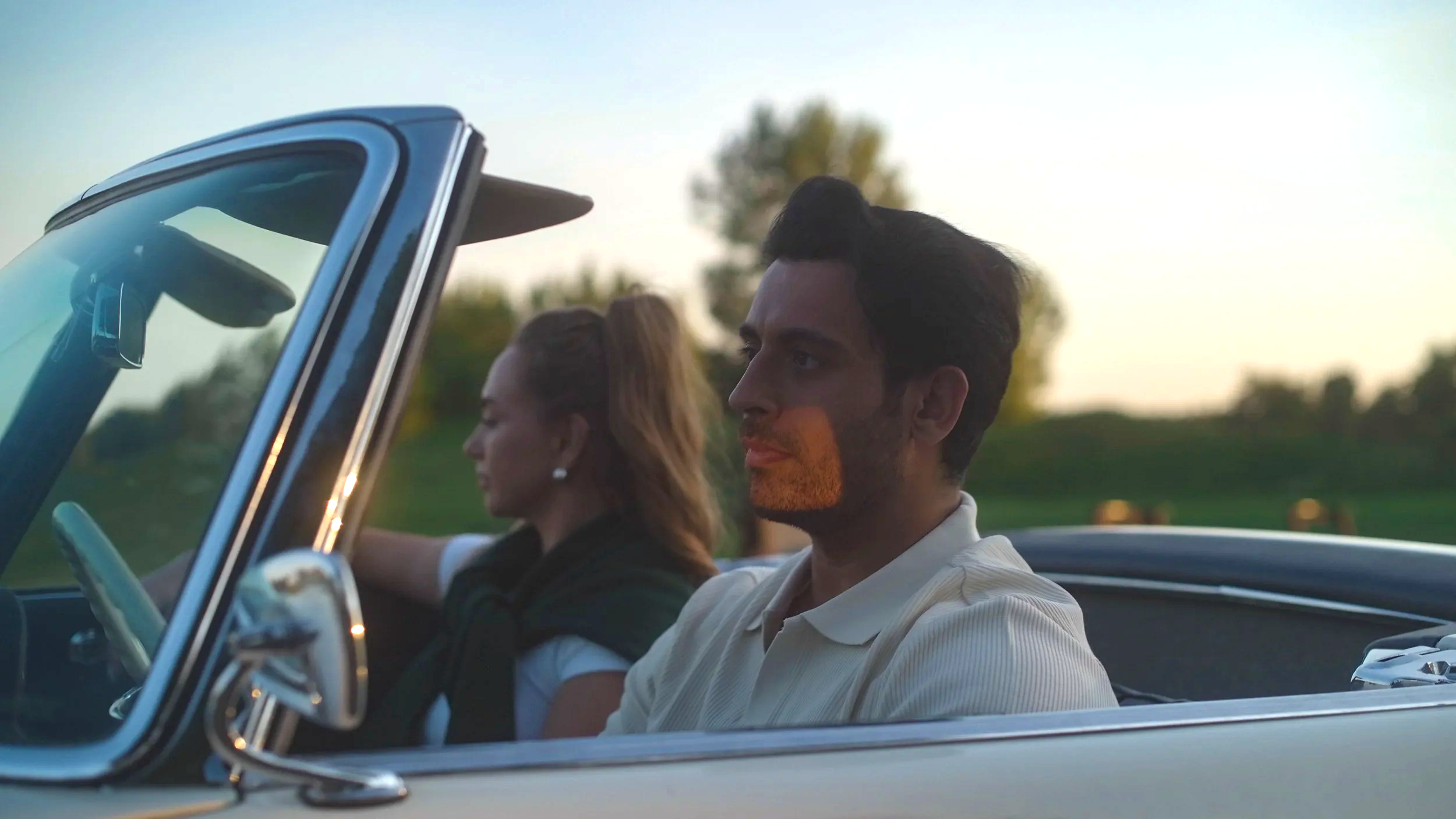A man and a woman driving in a convertible car on a country road during sunset, with trees and open fields in the background.
