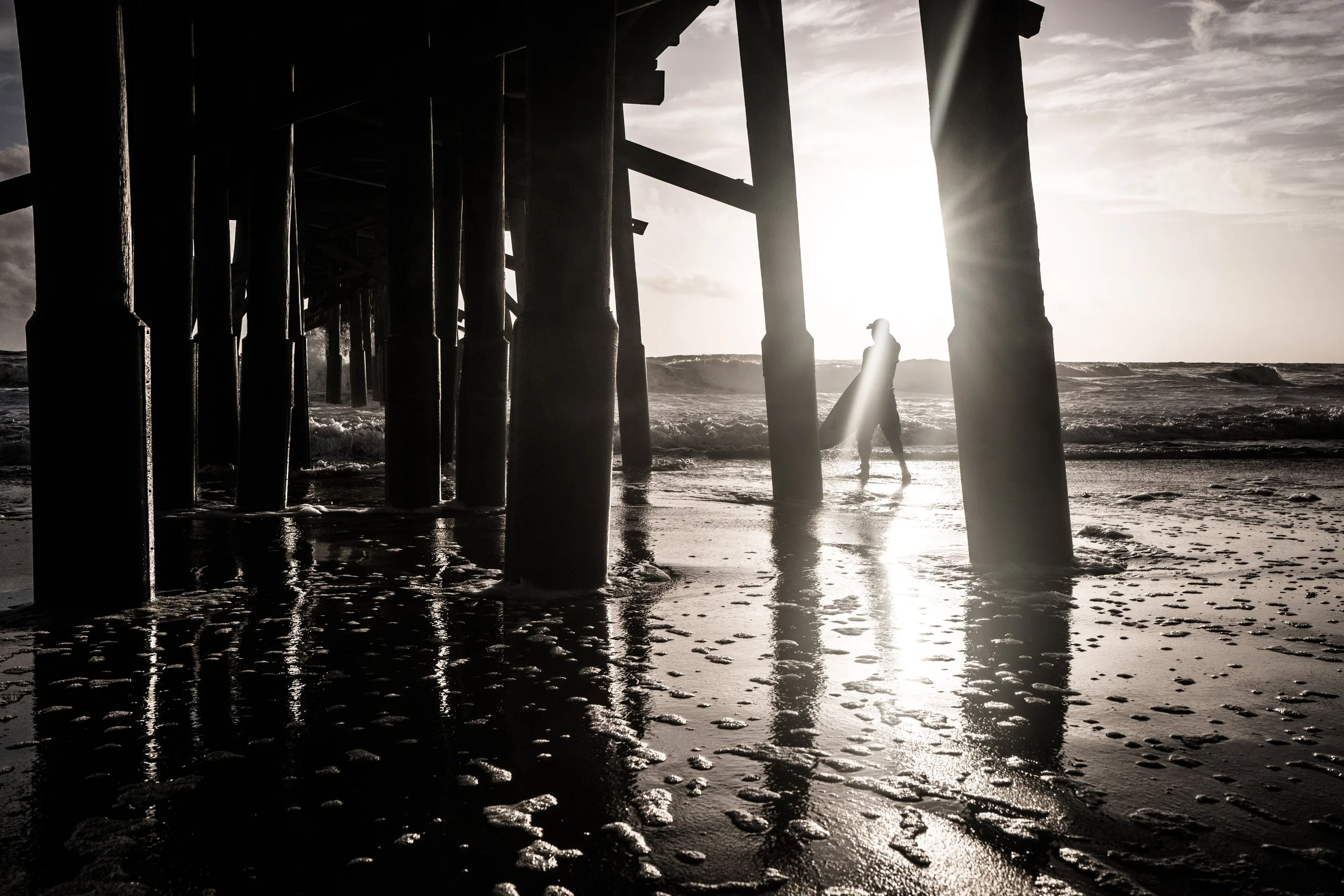 Coastal Vibes limited edition fine art photograph by Jason Sponseller, featuring a surfer walking beneath a beach pier in black and white coastal light, with ocean waves, wet sand, and dramatic shadow