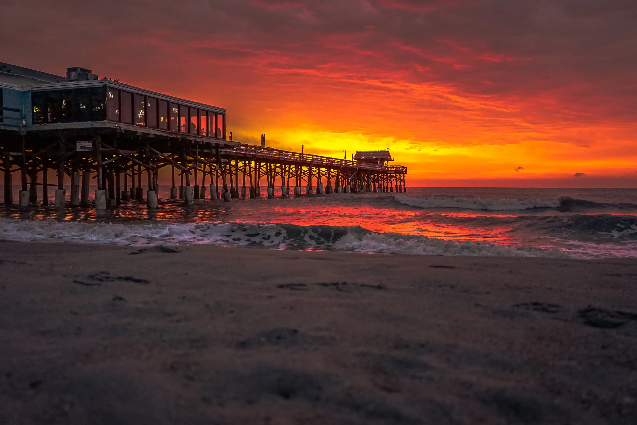 Coastal Vibes limited edition fine art photograph by Jason Sponseller, featuring Cocoa Beach Pier at sunrise with glowing red-orange skies, rolling ocean waves, and dramatic coastal light