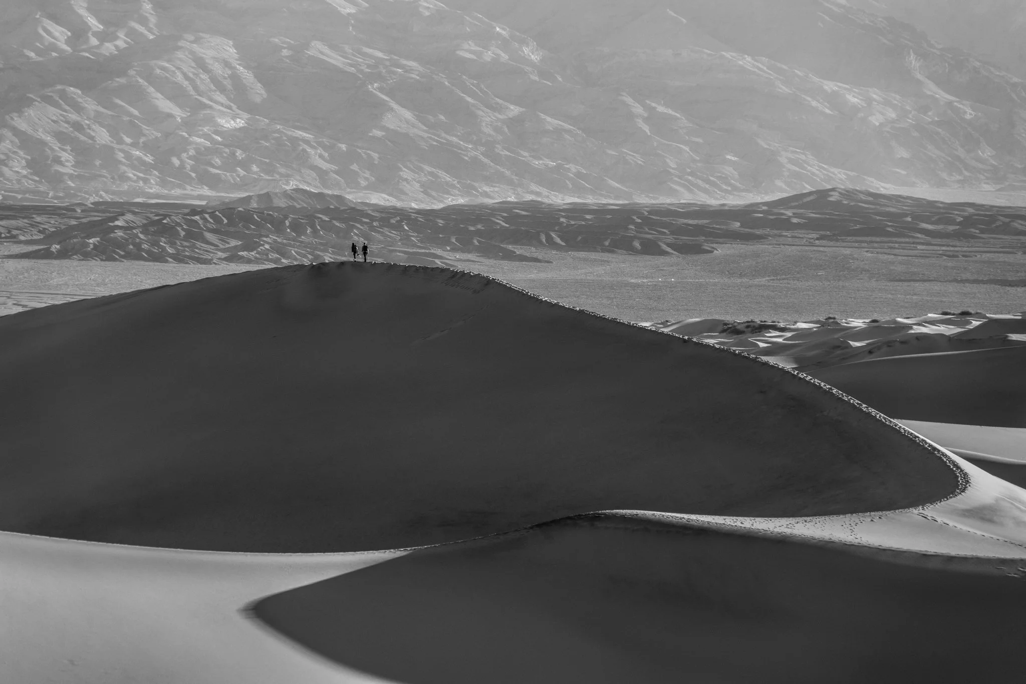Fine art black and white photograph of hikers atop the sand dunes in Death Valley, capturing an iconic desert landscape with sweeping textures, scale, and dramatic contrast