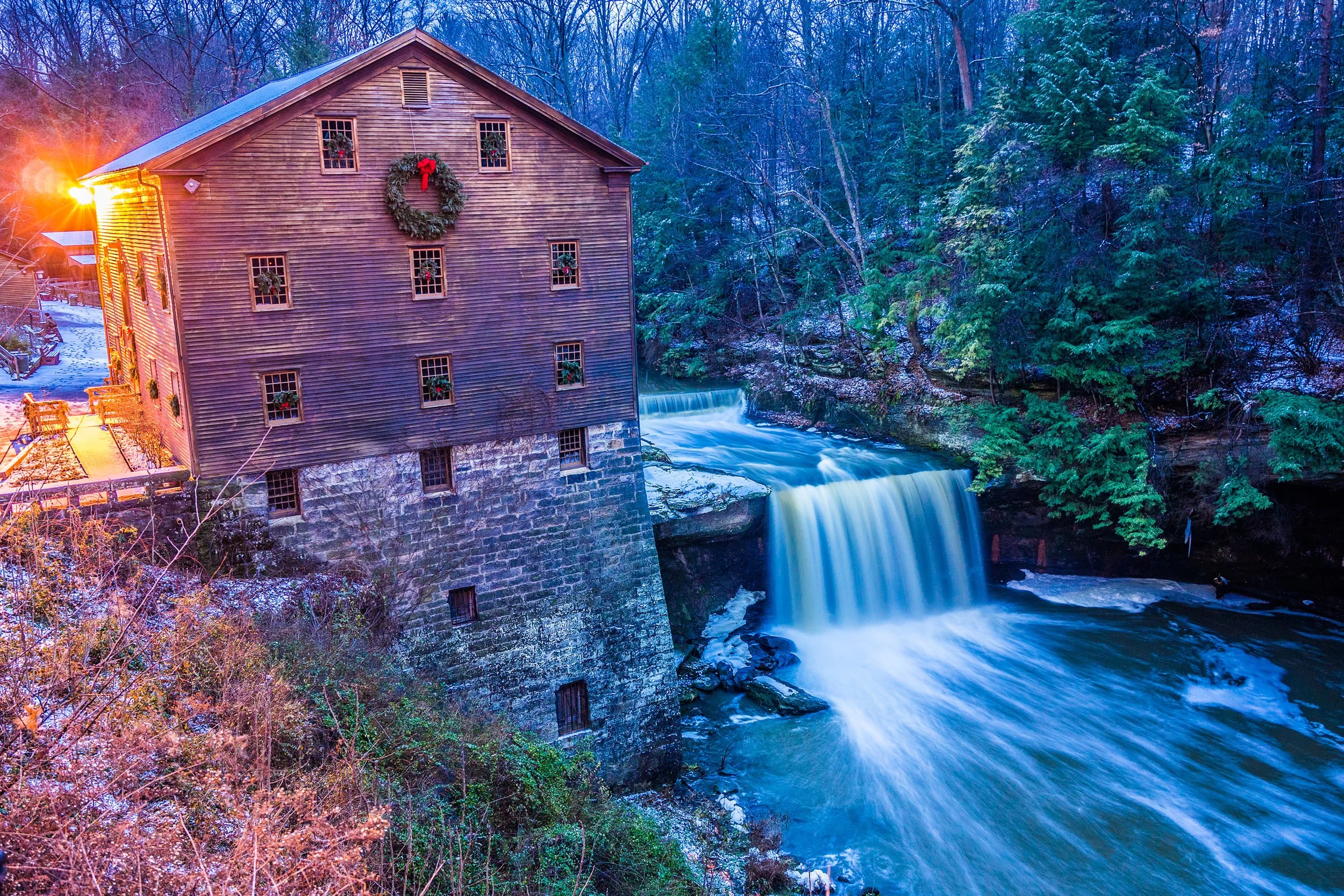 Dust and Distance: The Resonance of Time, limited edition fine art photograph by Jason Sponseller, featuring a historic watermill beside a waterfall in winter with flowing water, stone foundation, evergreen trees, and quiet old-world atmosphere