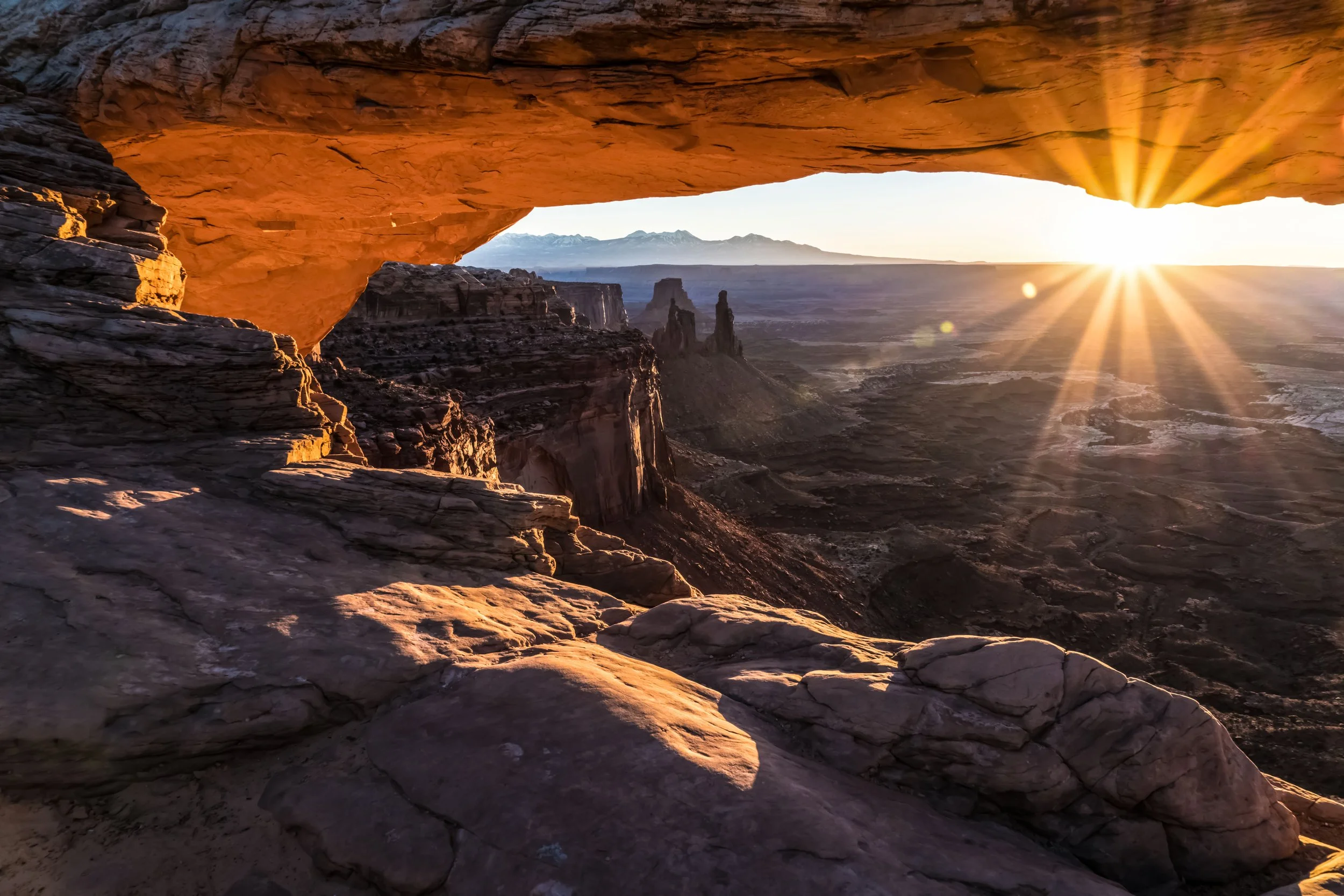 Dust and Distance: The Resonance of Time, limited edition fine art photograph by Jason Sponseller, featuring Mesa Arch in Canyonlands National Park at sunrise with golden desert light, red rock formations, canyon cliffs, and vast atmospheric distance