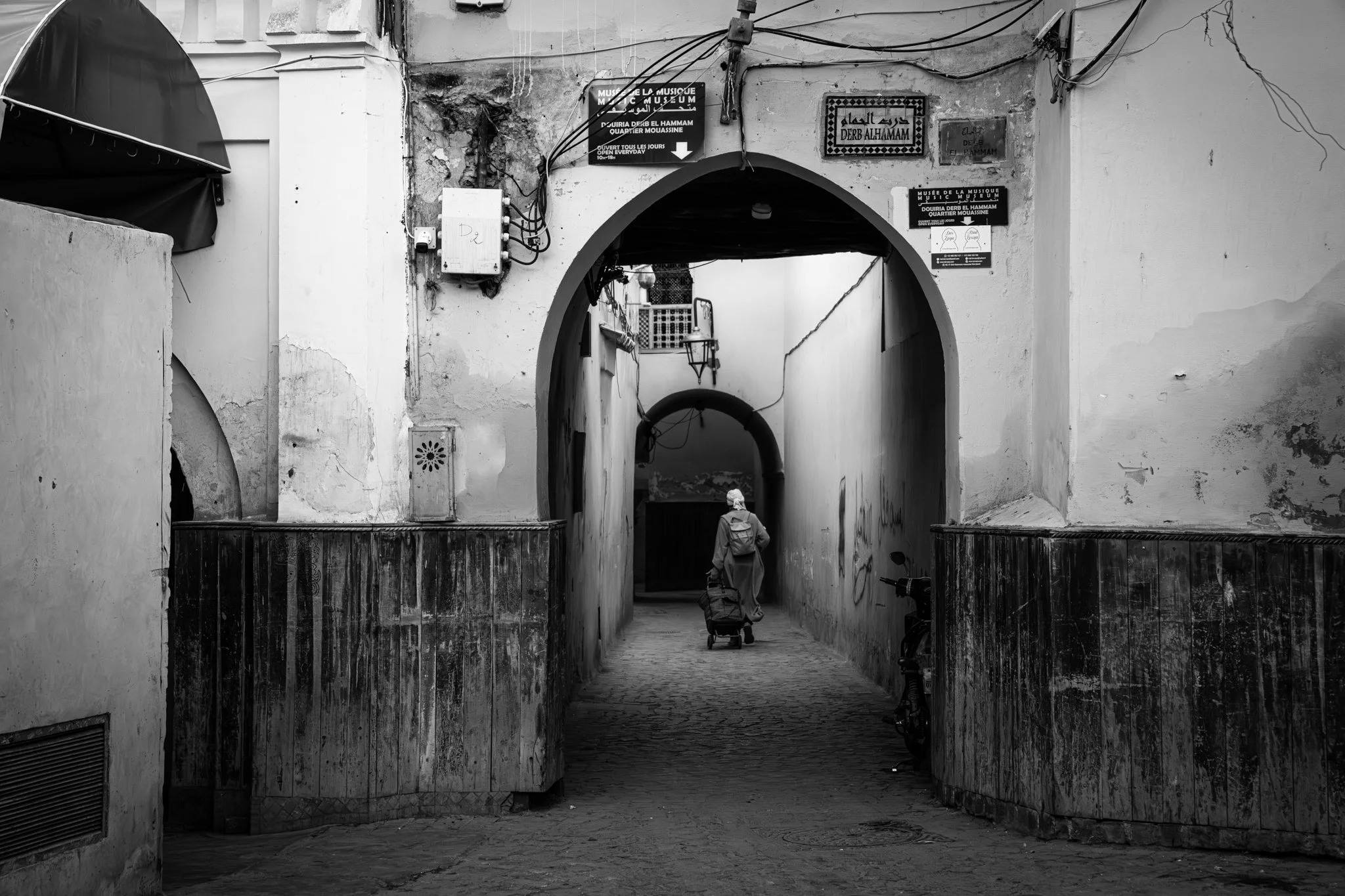 Echo Beneath the Stone, black-and-white fine art photograph by Jason Sponseller, featuring a lone figure walking through a Moroccan medina archway with historic architecture, layered shadow, and quiet human presence
