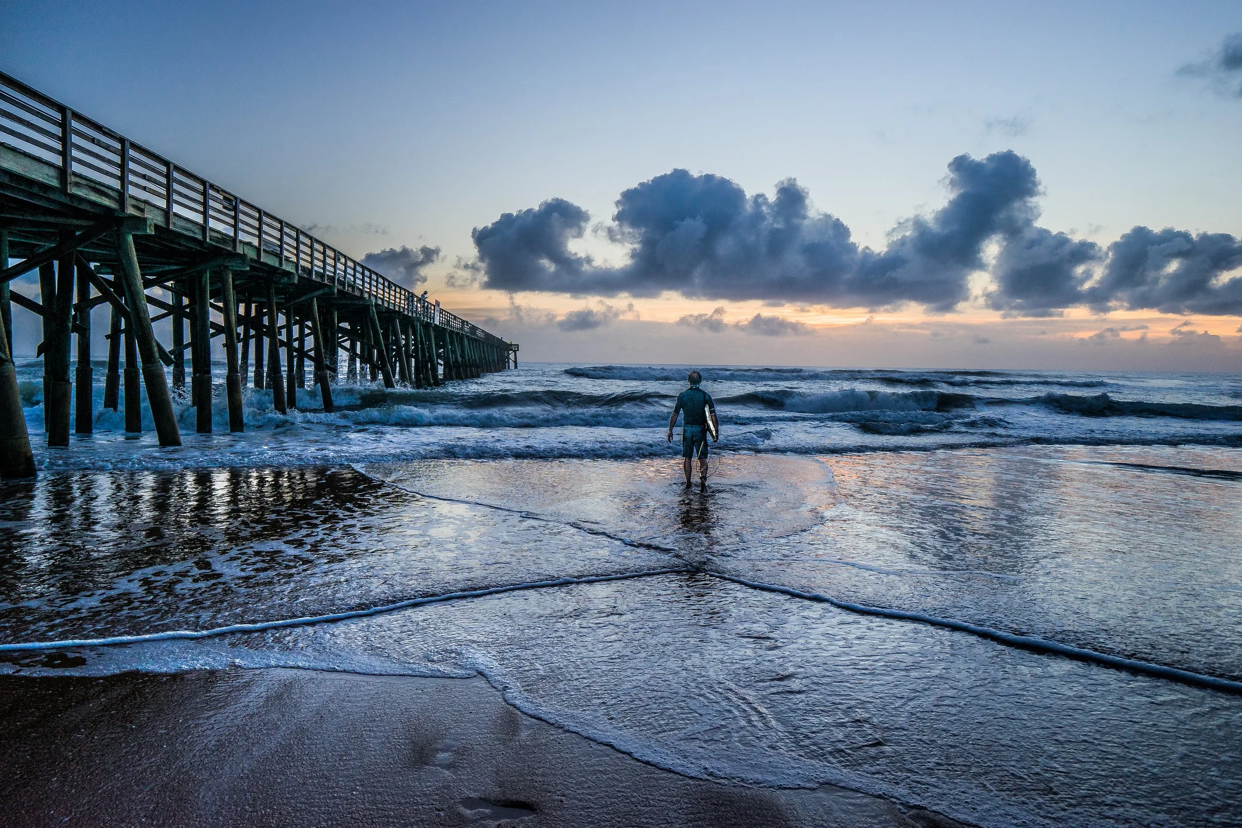 Coastal Vibes limited edition fine art photograph by Jason Sponseller, featuring a lone surfer walking toward the ocean beside St. Augustine Beach Pier at dawn with blue coastal light, waves, and dramatic clouds
