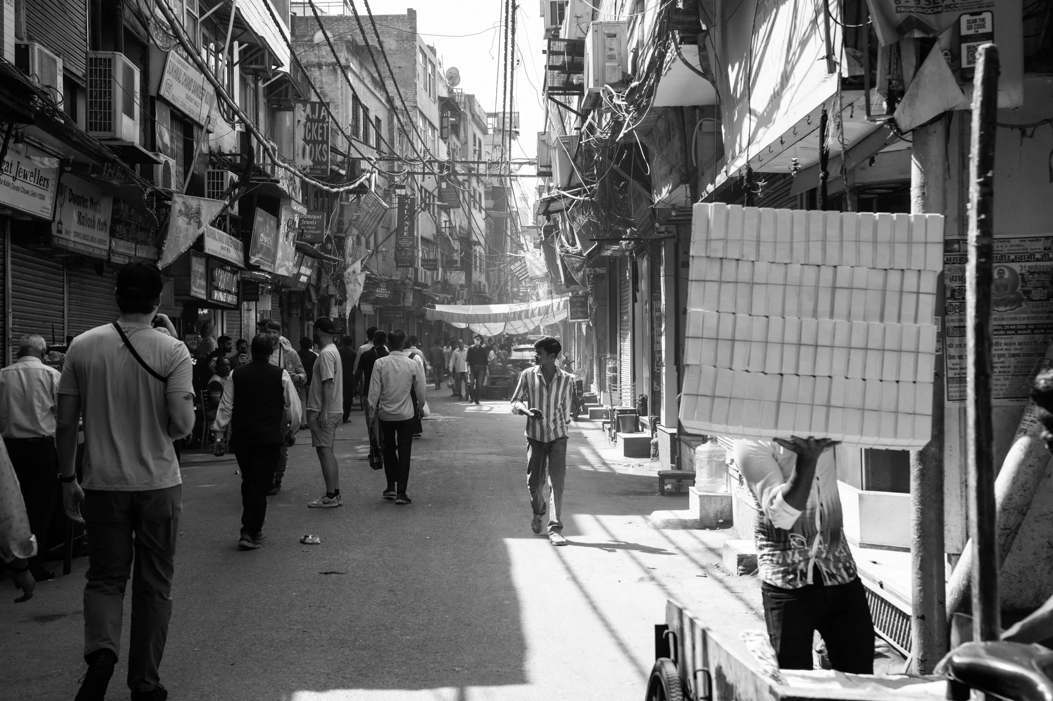 Echo Beneath the Stone, black-and-white documentary fine art photograph by Jason Sponseller, featuring a busy India market street with layered architecture, overhead wires, shadow, and human presence